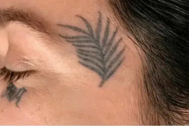 Close-up of a person's face; a gray palm leaf tattoo is on their cheek near the eye.