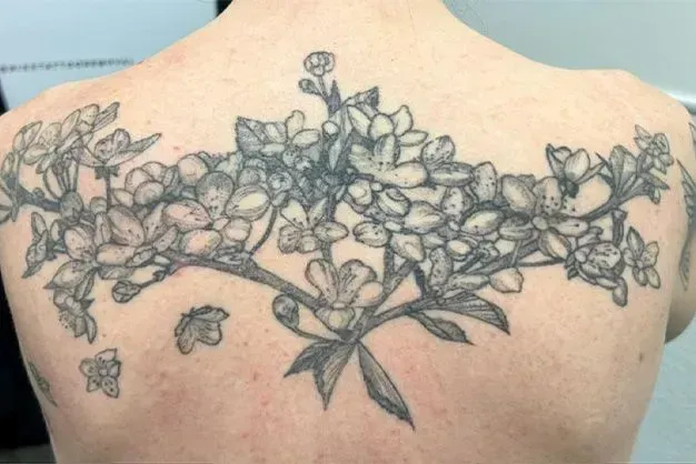 Back tattoo of a blossoming floral branch in black and gray ink.