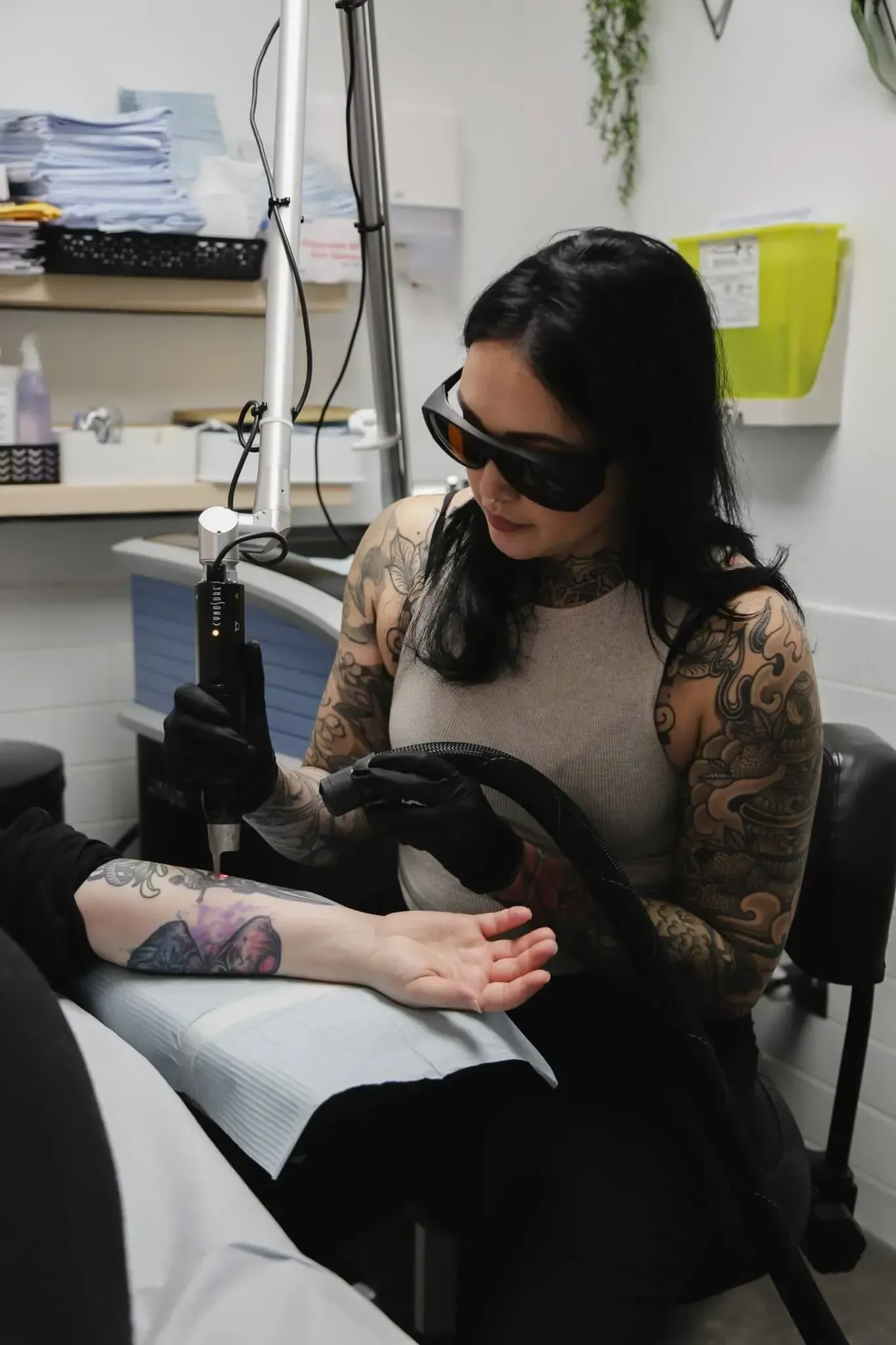 A technician with tattooed arms uses a laser device to remove a tattoo from a client's arm in a clinical setting.