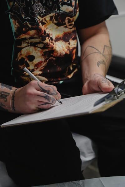 Person with tattoos on arms drawing on clipboard, wearing a black shirt.