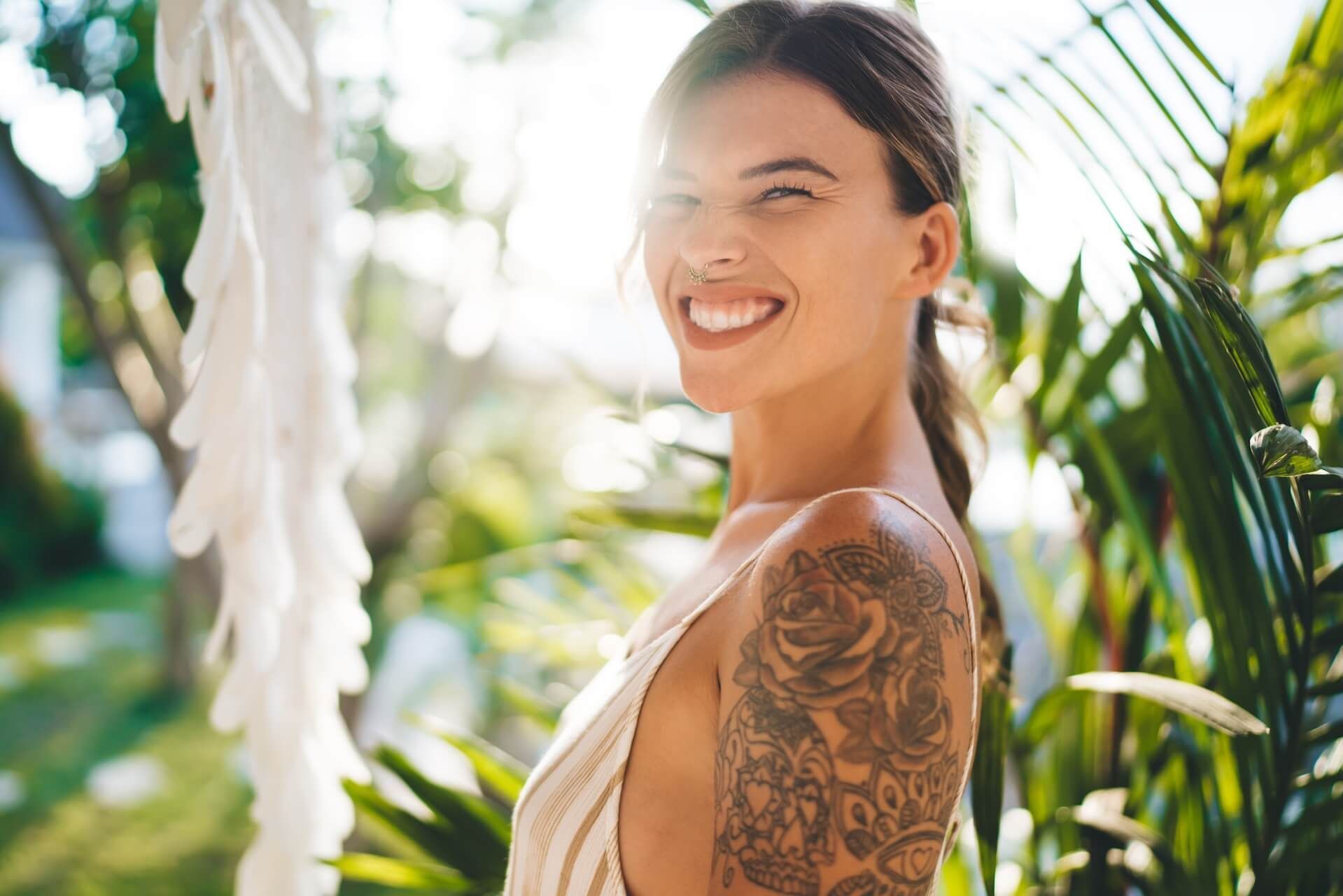 Smiling woman with tattooed arm standing among sunlit tropical plants