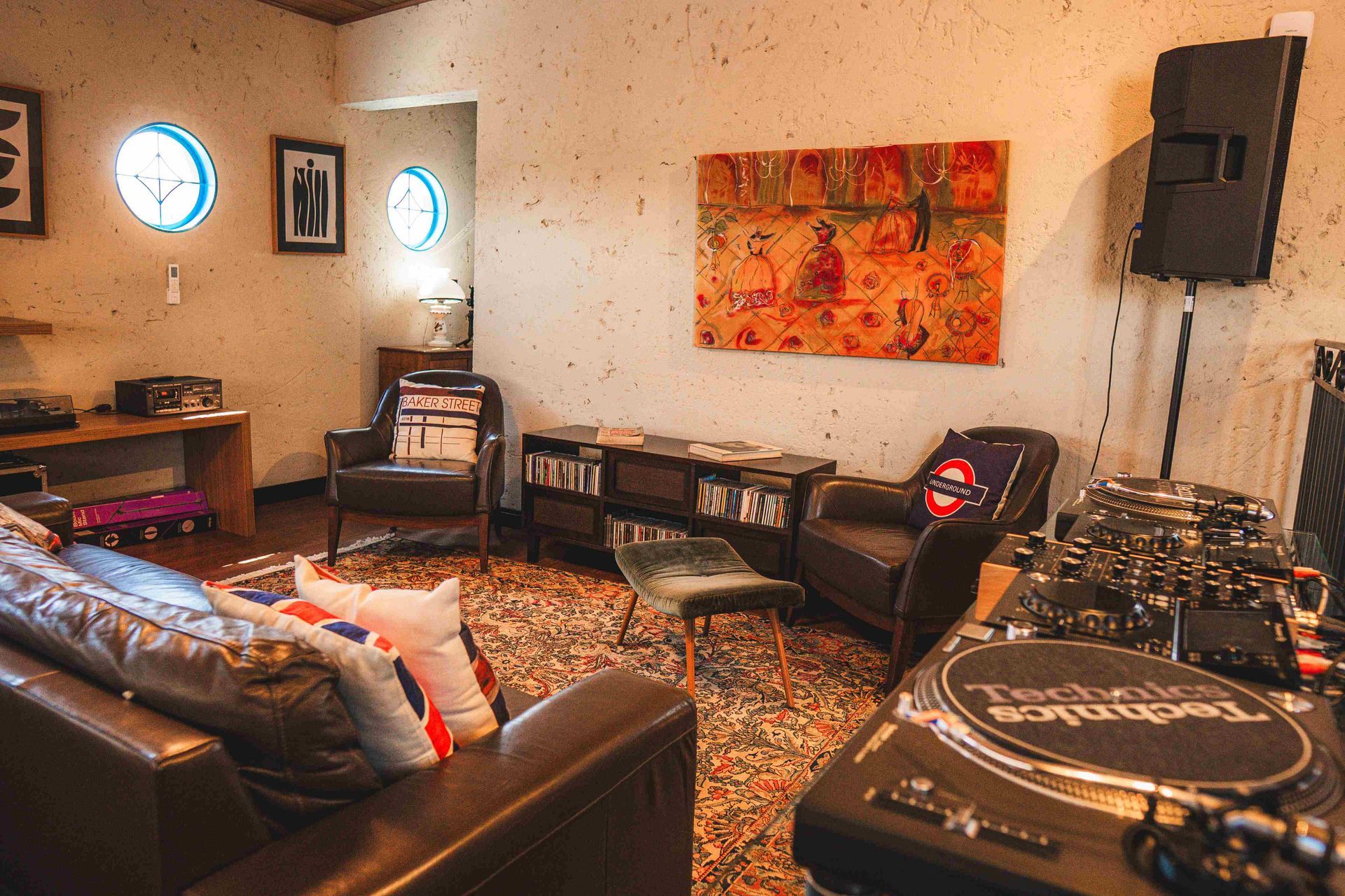 Uma sala de estar com um sofá de couro, duas poltronas, um tapete estampado, um quadro na parede e equipamento de DJ sobre uma mesa.