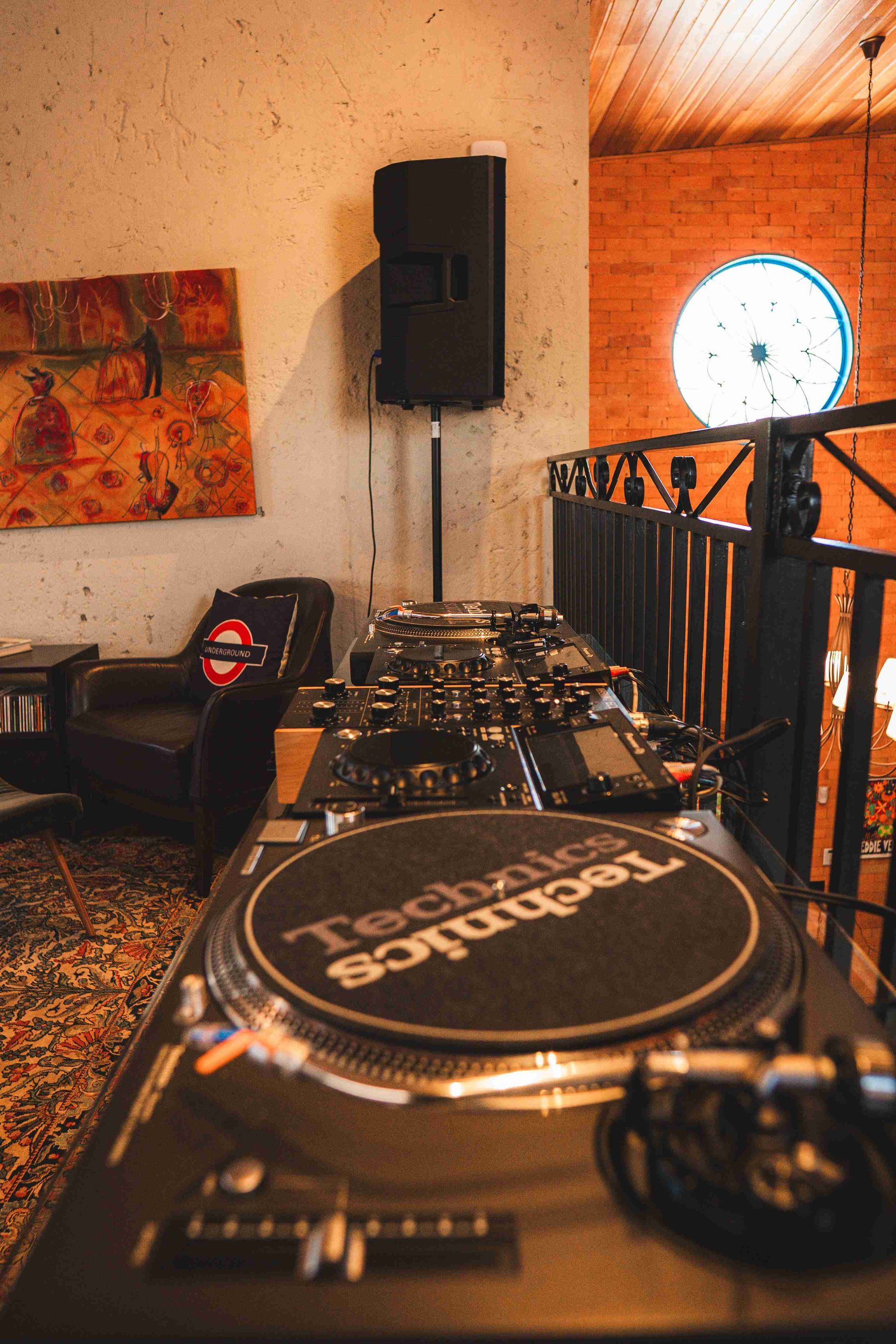 Uma cabine de DJ com um toca-discos Technics em primeiro plano, situada em uma sala interna com uma caixa de som e uma poltrona escura.
