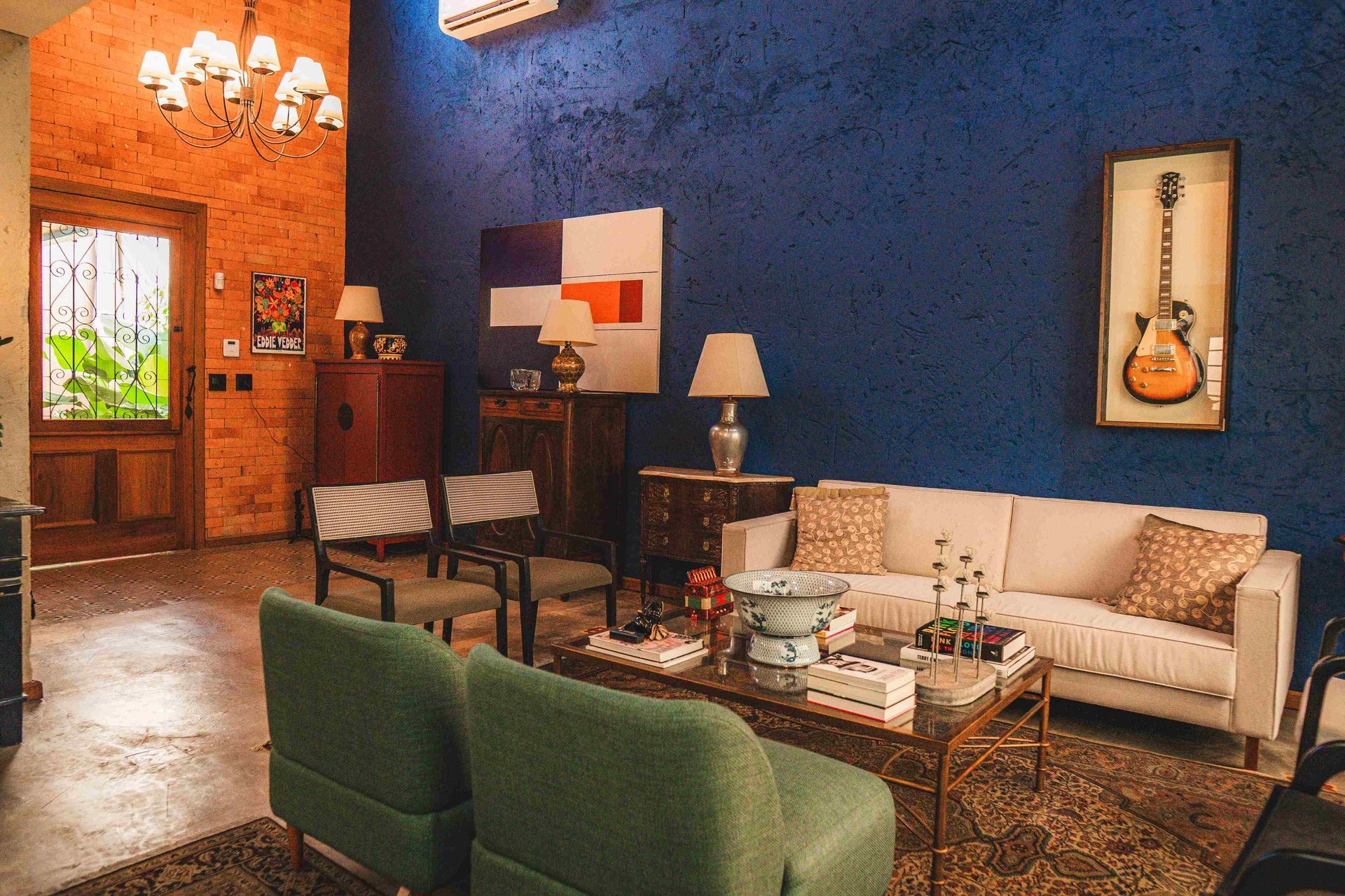 Uma sala de estar com paredes azuis, um sofá branco, duas poltronas verdes, uma mesa de centro e um violão pendurado na parede.
