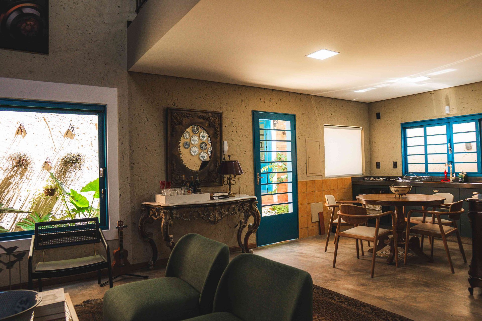 Uma sala de estar bem iluminada com duas poltronas verdes, uma mesa de jantar de madeira e janelas e portas com molduras azuis.