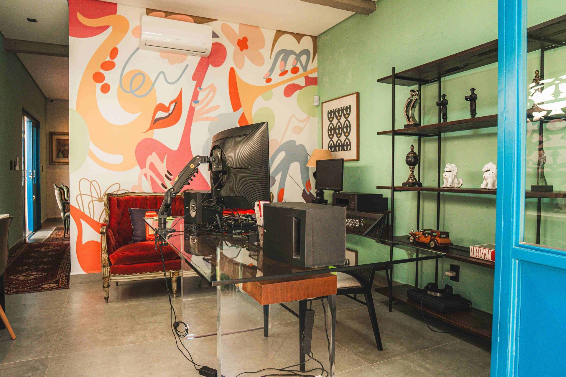 Um escritório em casa com uma mesa de vidro, uma cadeira de veludo vermelho, arte abstrata na parede e uma estante em níveis contra uma parede verde.