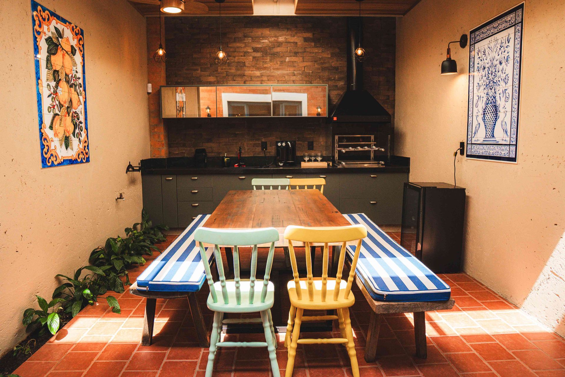 Uma cozinha e sala de jantar internas com mesa de madeira, cadeiras coloridas, bancos listrados e piso de tijolos.