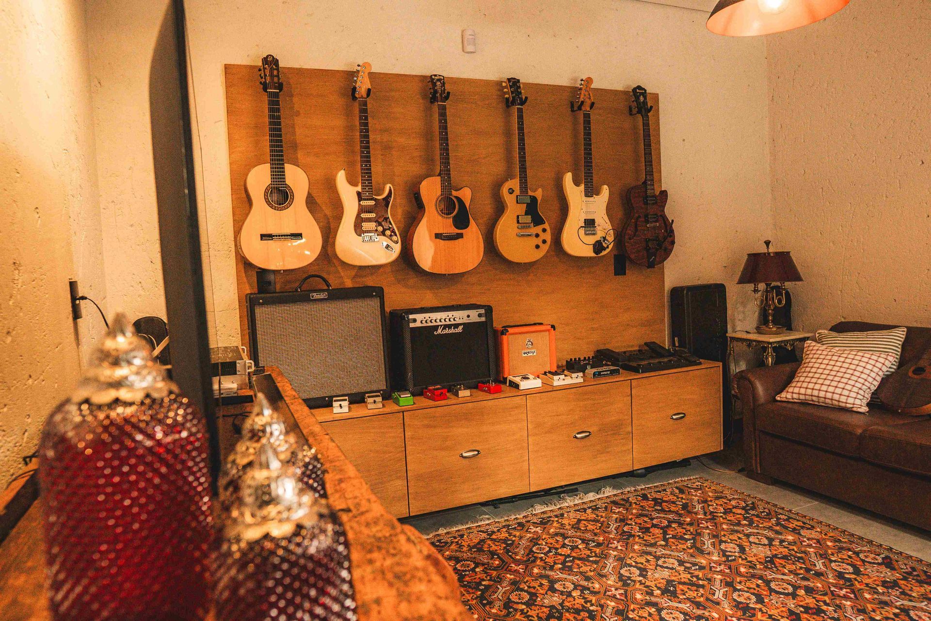 Um estúdio de música com uma fileira de seis guitarras penduradas em uma parede de madeira, acima de amplificadores e um sofá de couro.
