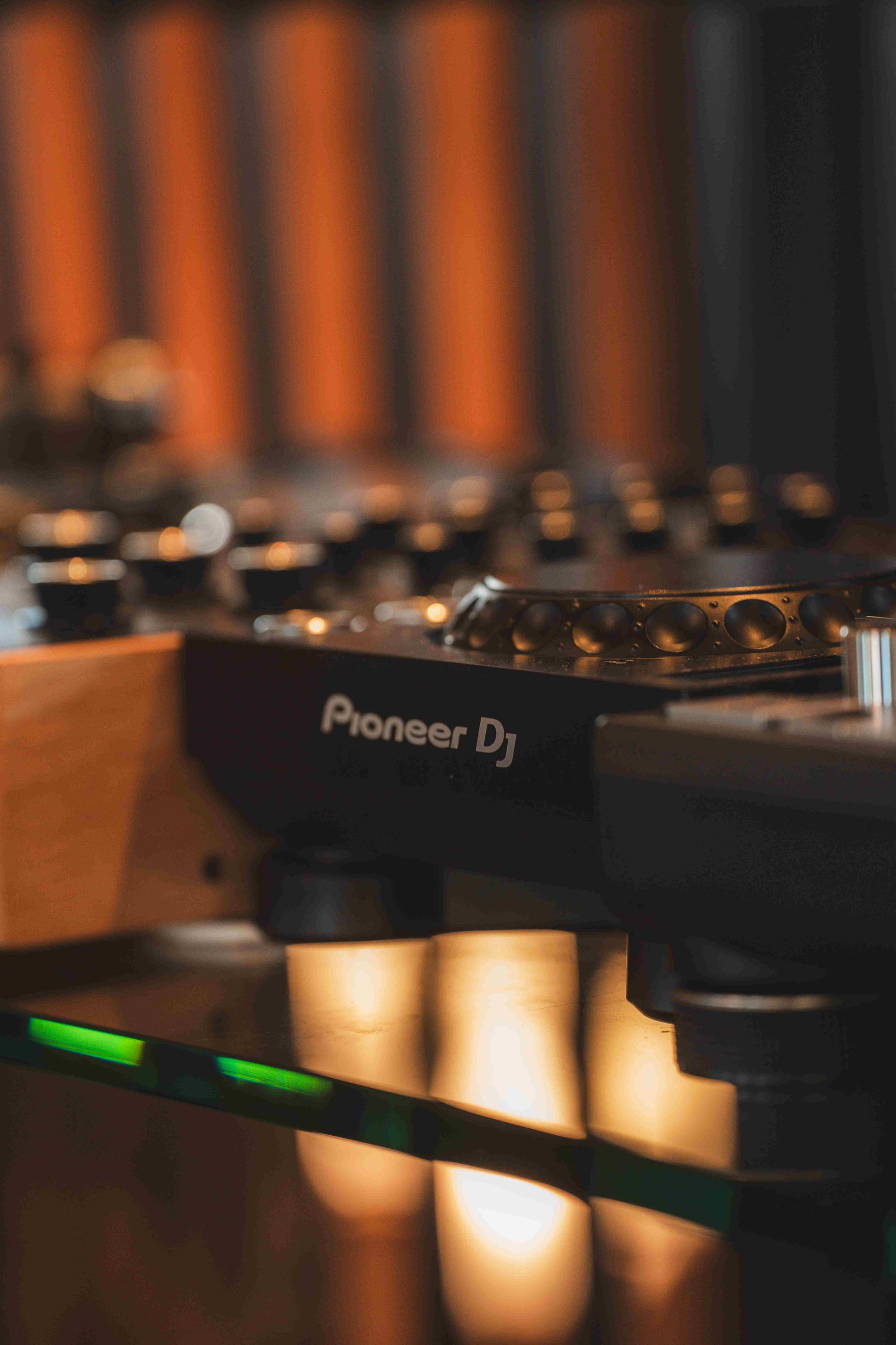 Vista lateral aproximada de um controlador Pioneer DJ preto apoiado em uma superfície refletora com luzes quentes e brilhantes.