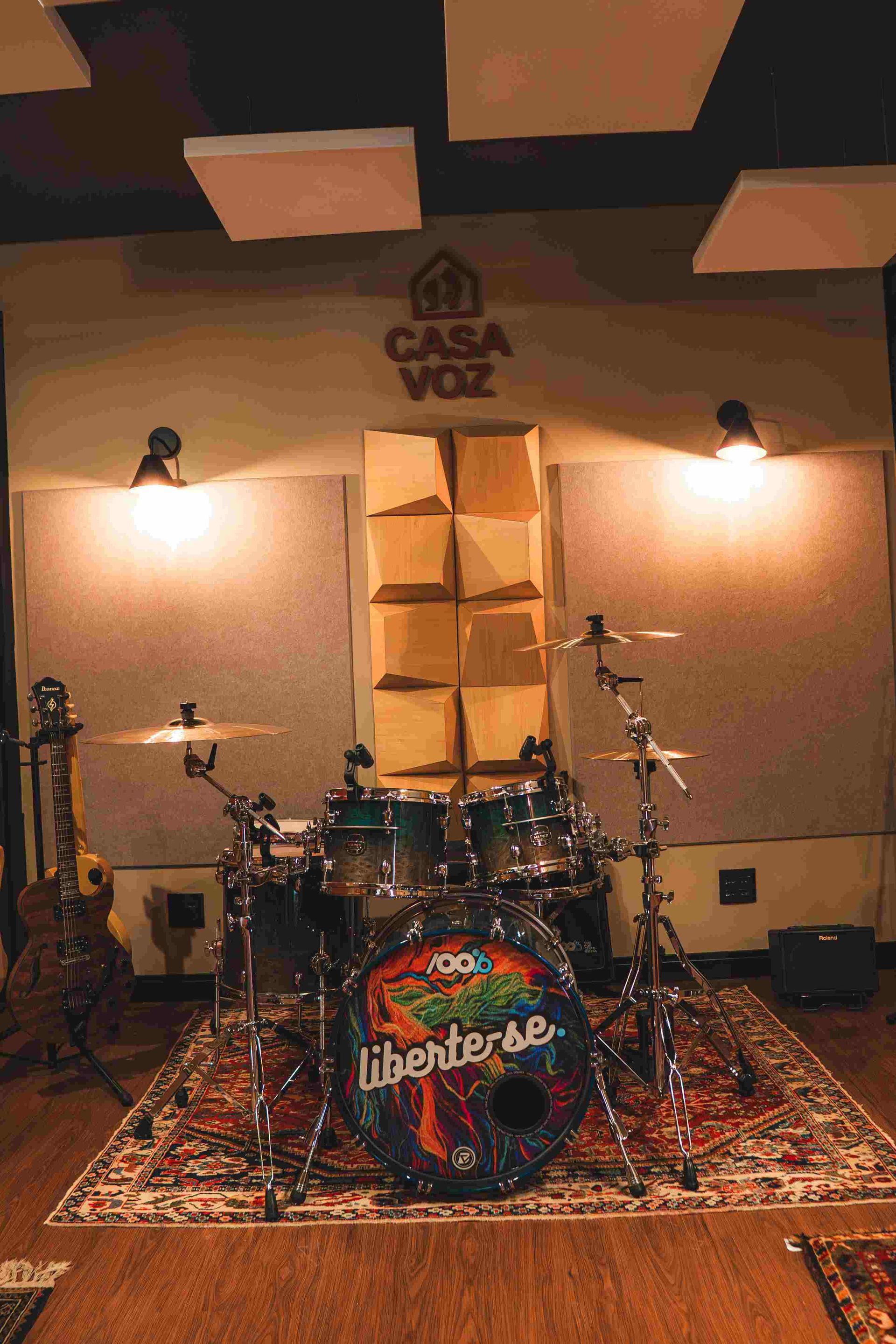 Uma bateria com um logotipo colorido no bumbo está sobre um tapete ornamentado em um estúdio de música com painéis de parede à prova de som.