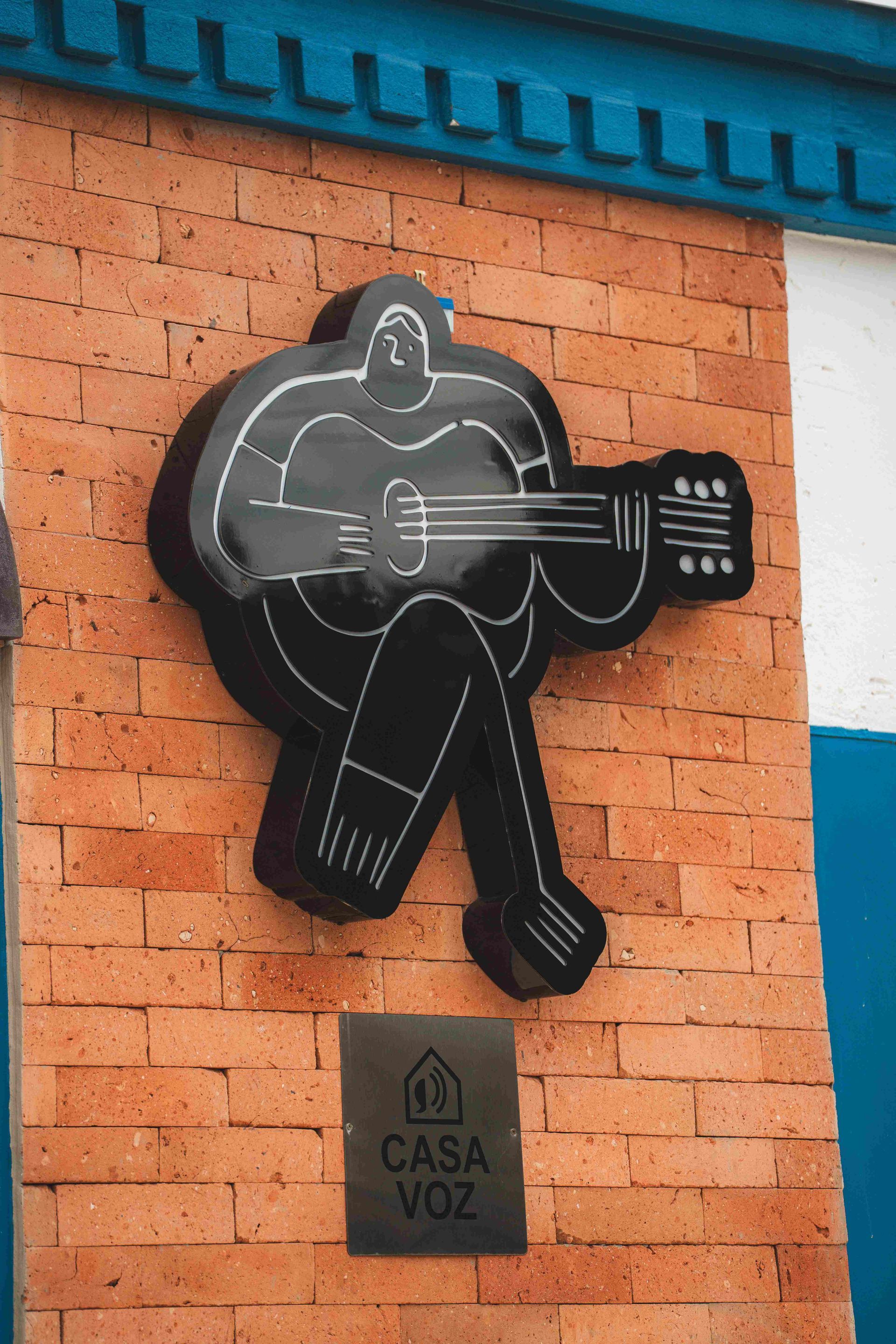 Uma figura estilizada em metal preto, recortada de uma pessoa tocando guitarra, está fixada em uma parede de tijolos acima de uma placa com os dizeres 