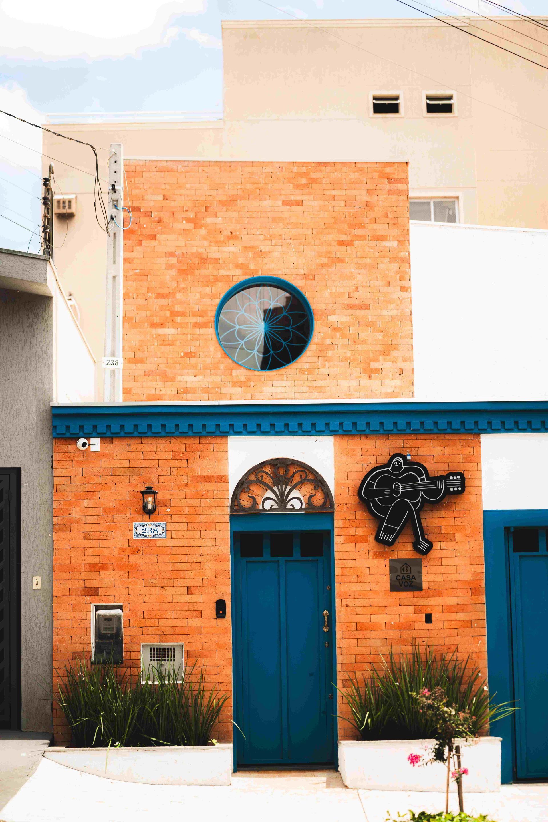 A fachada de um prédio de tijolos apresenta uma porta azul em arco, uma janela redonda e uma escultura de parede representando uma constelação, ladeada por plantas.