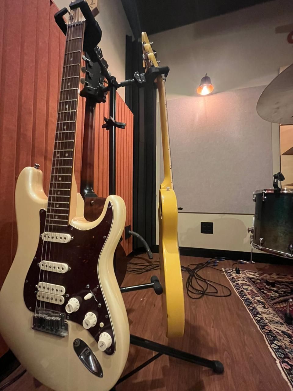 Duas guitarras elétricas em um suporte em um estúdio de música, com uma bateria visível ao fundo.