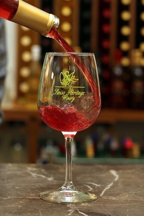 Red wine being poured into a clear glass with a winery logo; blurred background of shelves.