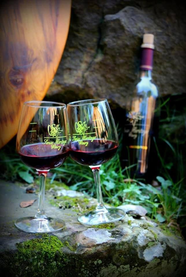 Two wine glasses filled with red wine, bottle of wine, sitting on mossy stone, in front of a wooden object and rocks.
