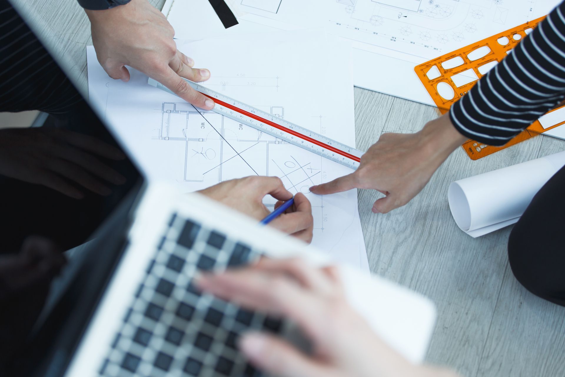 Hands working on architectural plans with ruler and laptop.