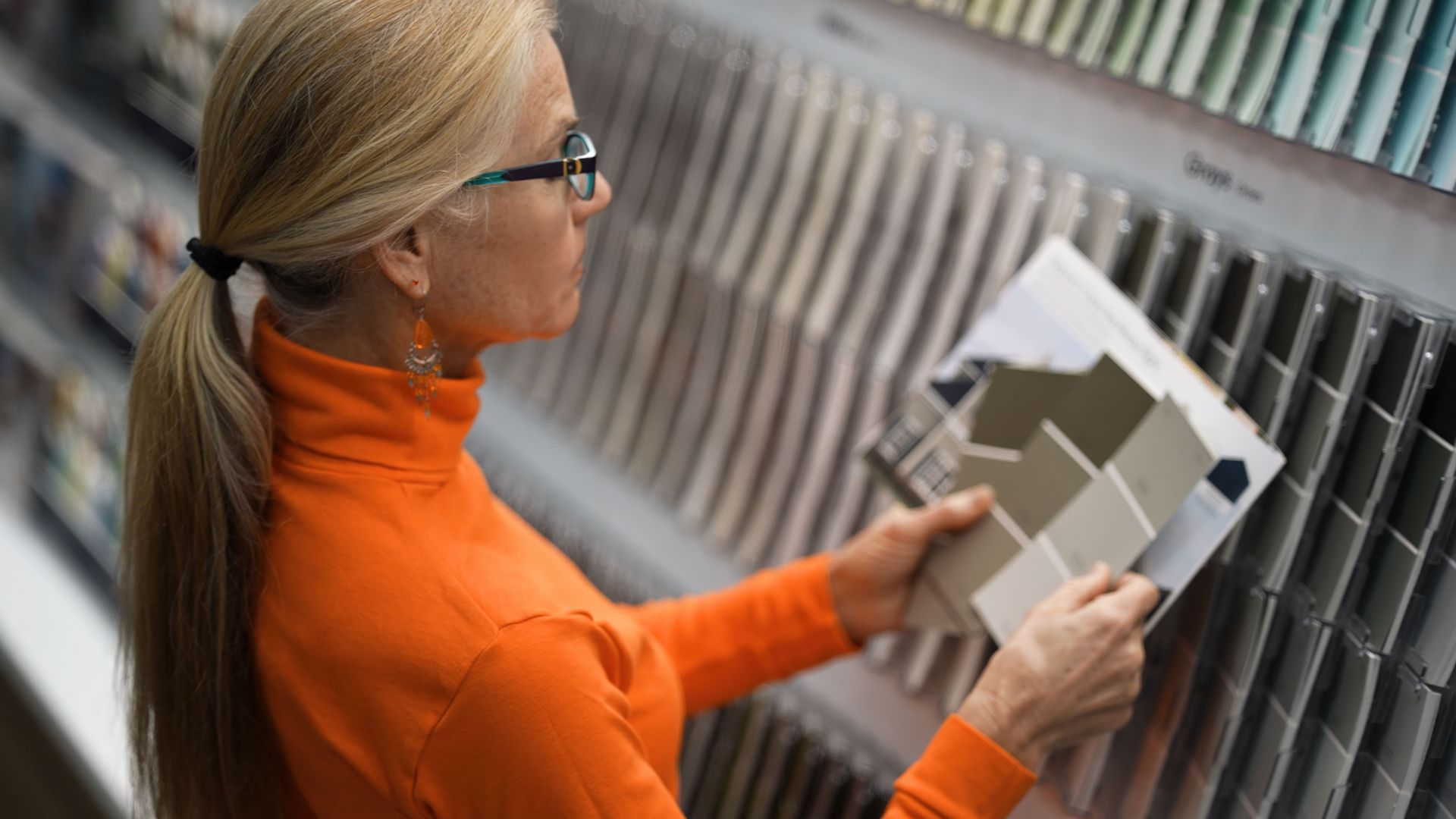 Woman with glasses, orange top, looking at paint swatches in a store.
