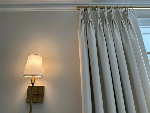 Off-white curtains with pinch pleats hang next to a wall-mounted brass lamp on a cream-colored wall.