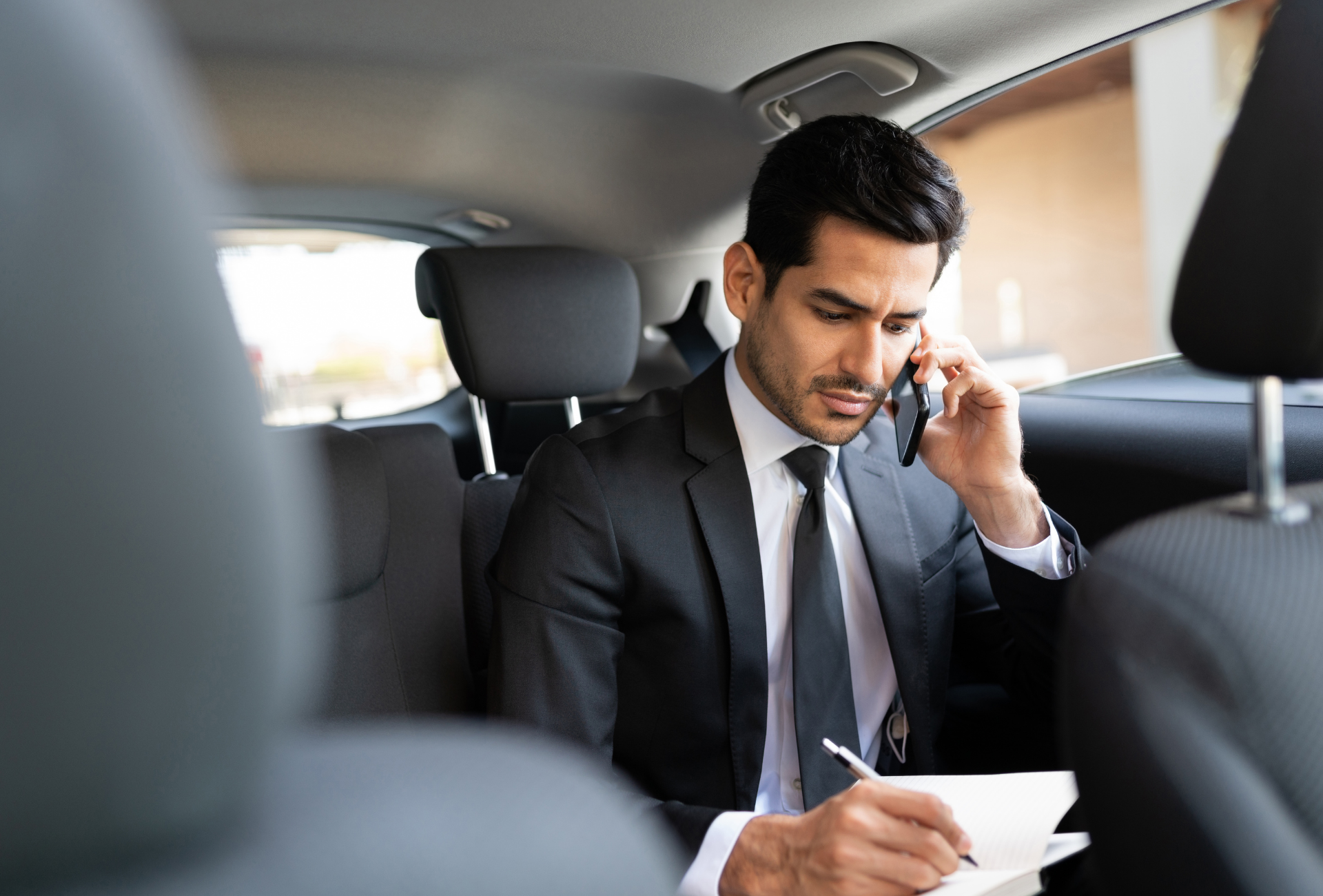 A man in a suit and tie is sitting in the back seat of a car talking on a cell phone.