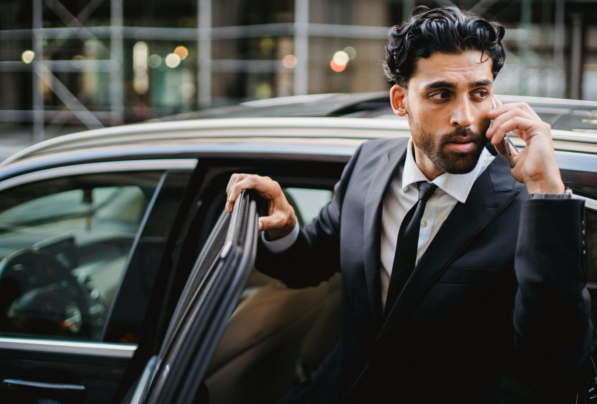 A man in a suit and tie is talking on a cell phone while getting out of a car.