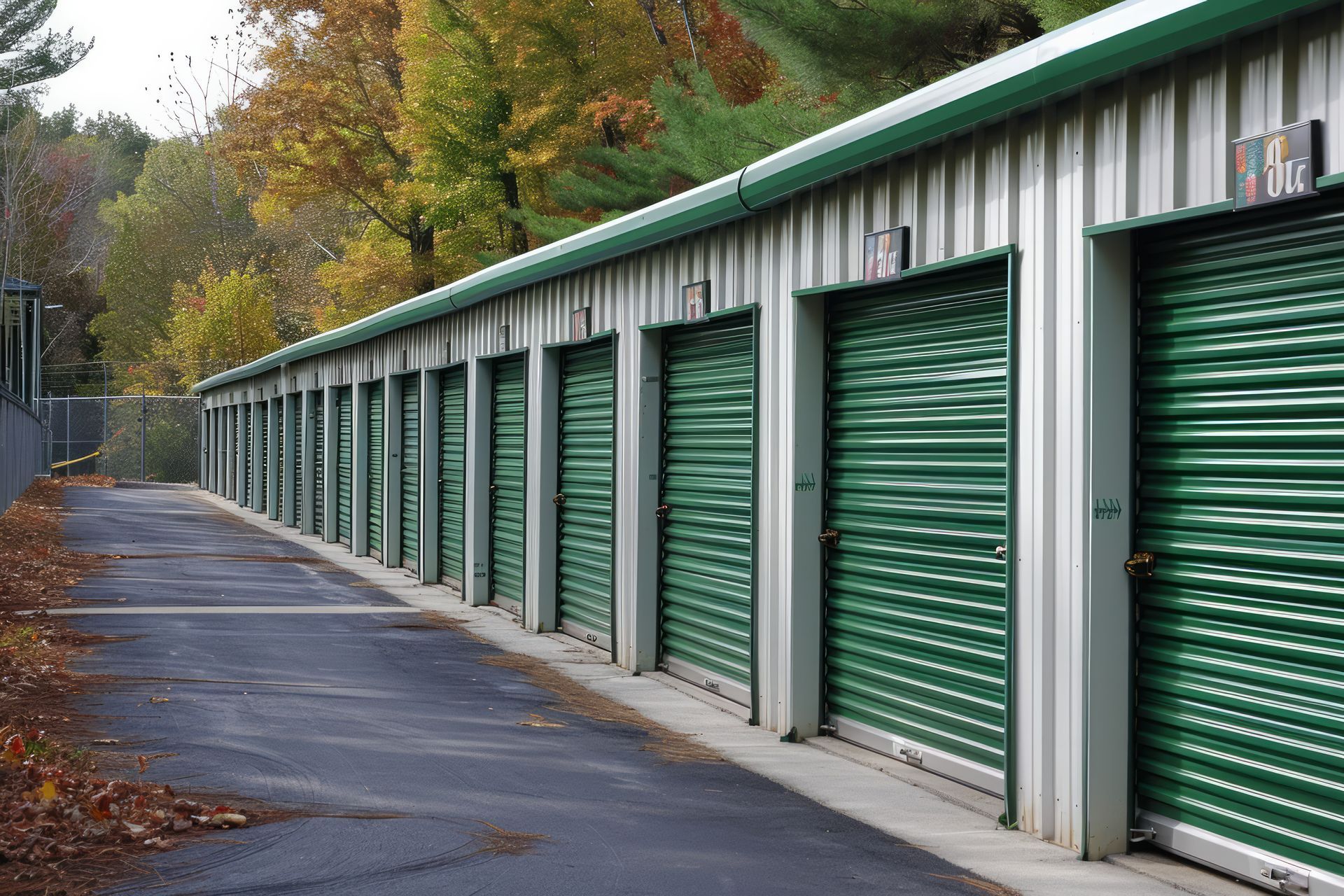 Row of green storage unit doors along an outdoor driveway beside autumn trees.