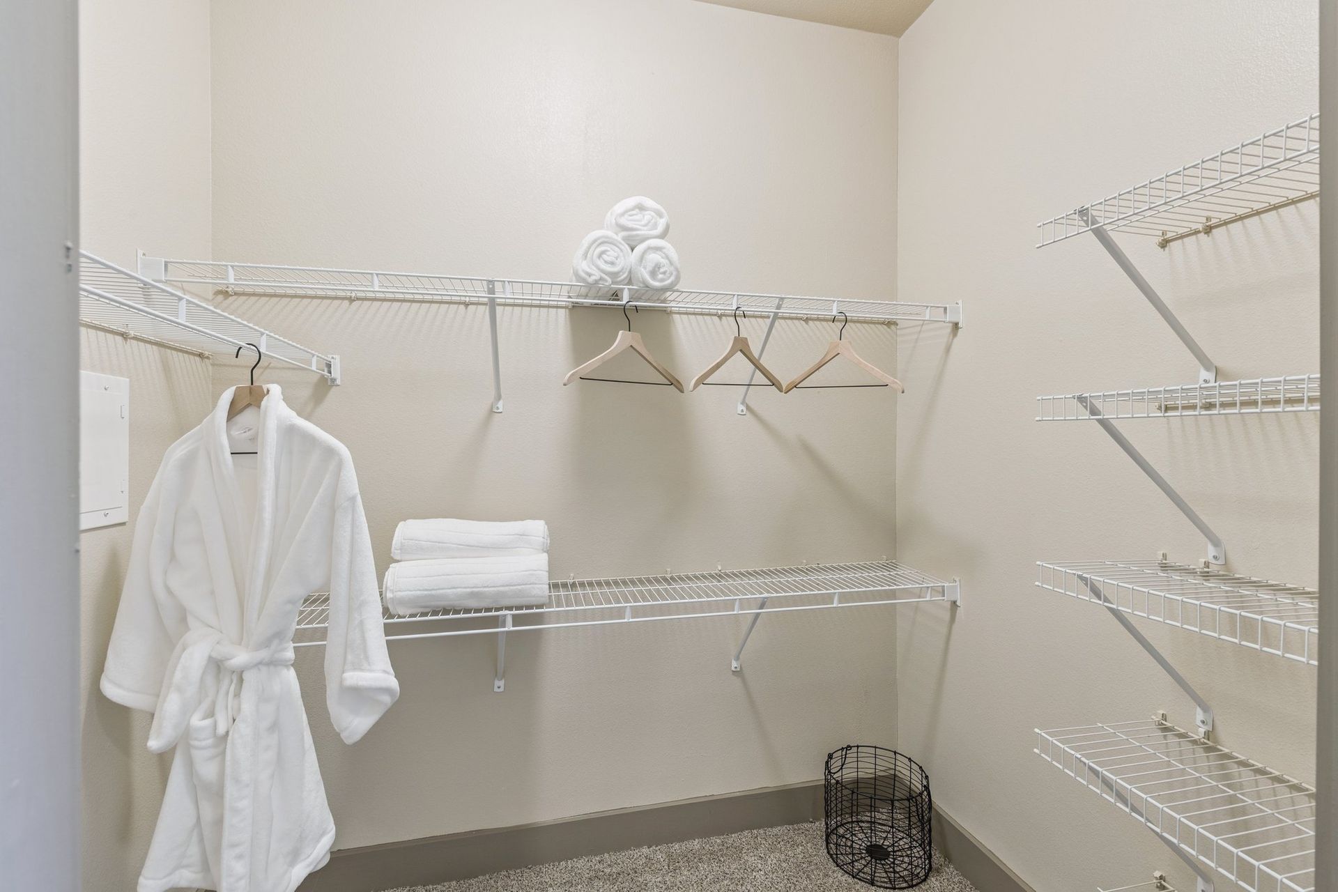 Empty walk-in closet with wire shelving. A white robe hangs on the left. White towels are stacked and rolled on shelves.