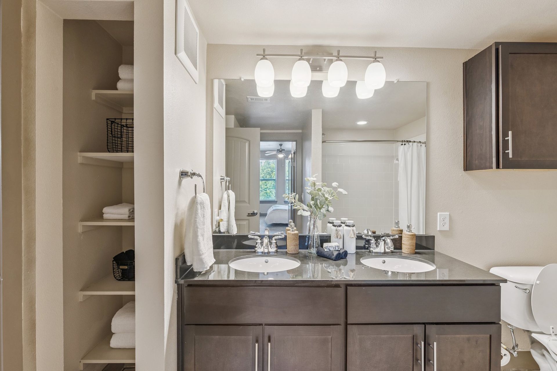 Bathroom with double sink vanity, dark cabinets, bright lighting, and built-in shelving with towels.