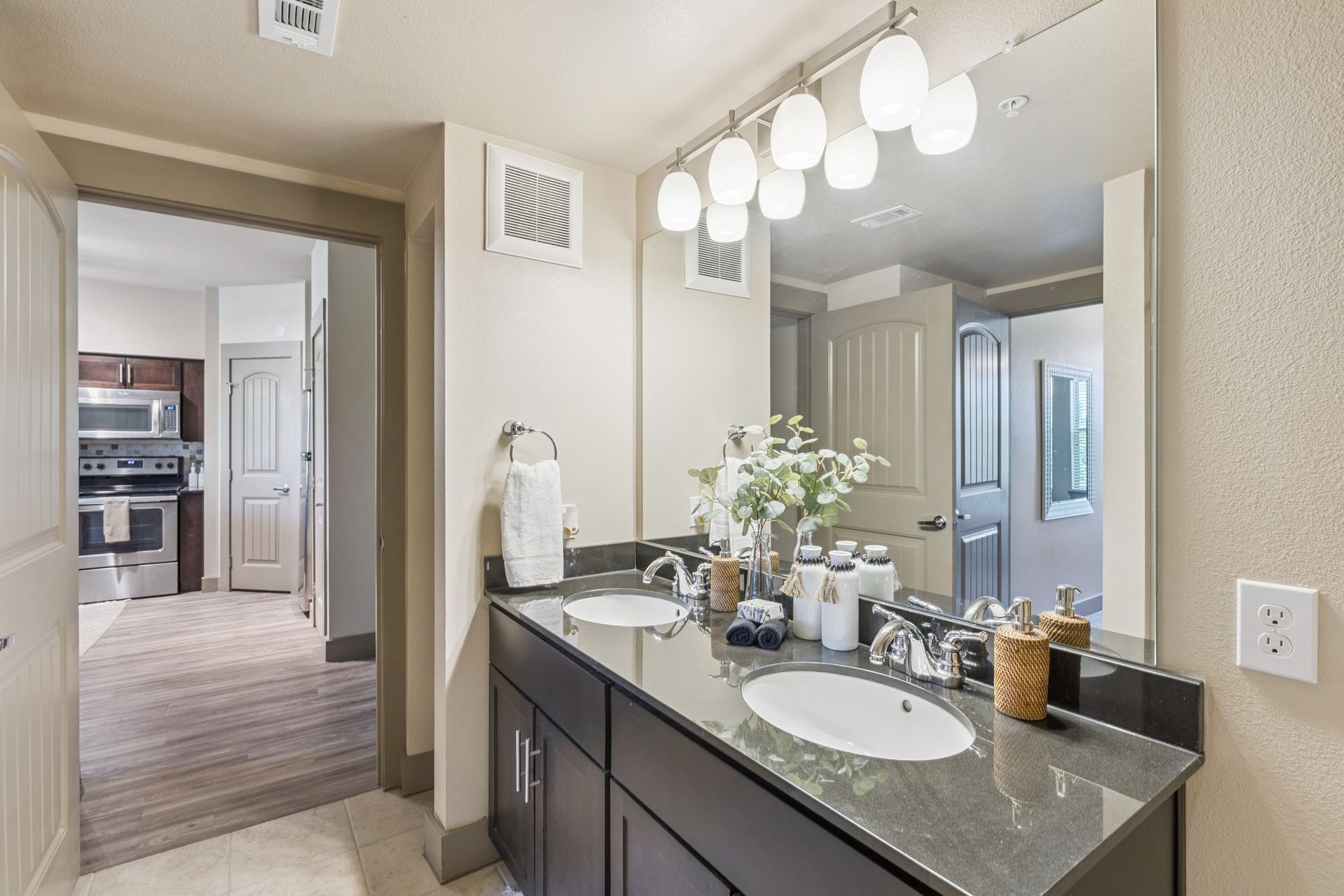 Bathroom with double sinks, large mirror, and overhead lighting; view into the kitchen.