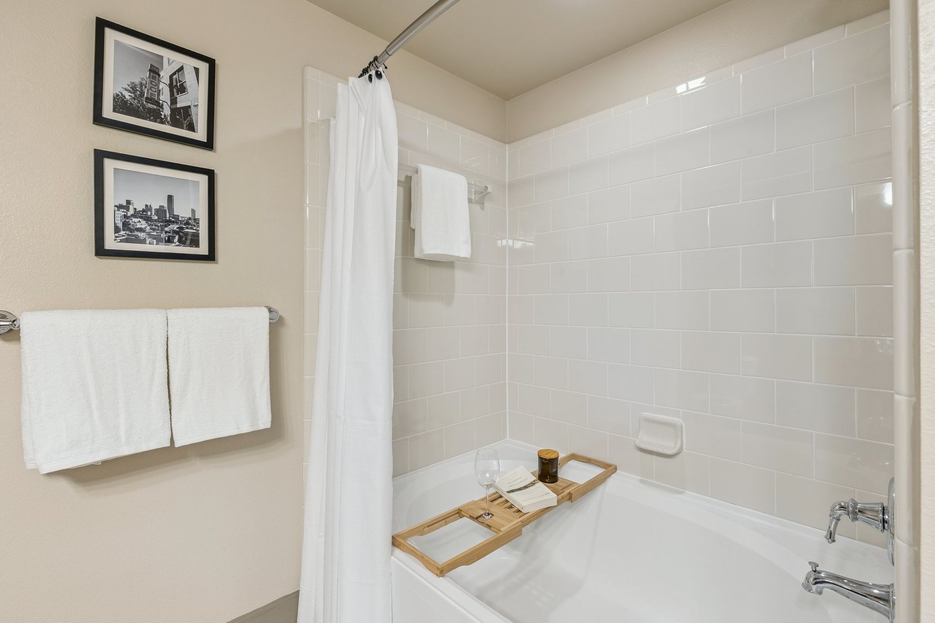 Bathroom with white tub, white tile, and tan walls. Includes a white shower curtain and two framed pictures.