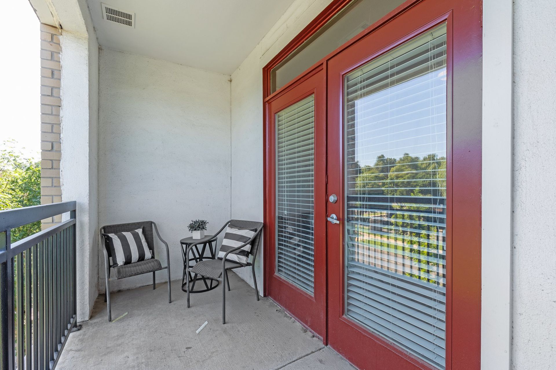 Balcony with two chairs, small table, and red-framed glass doors. Blinds, metal railing, and brick column.