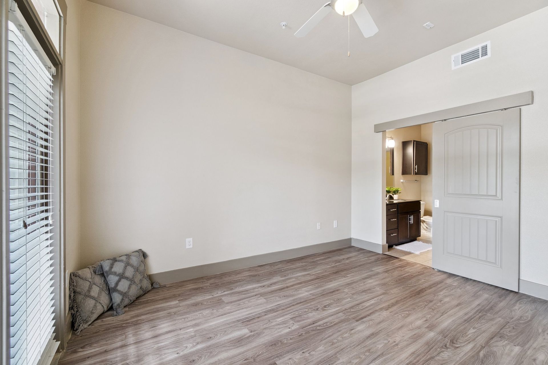 Empty bedroom with wood flooring, large window, and sliding door to bathroom.