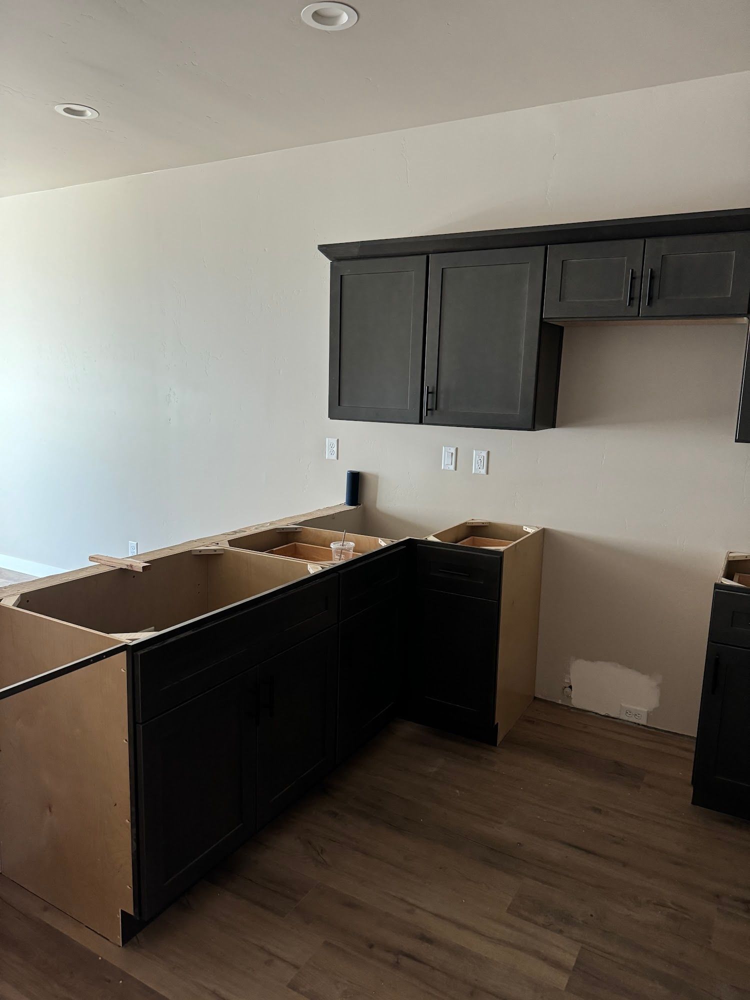 Kitchen cabinets in various stages of installation: dark lower cabinets, upper cabinets, unfinished walls, wood flooring.