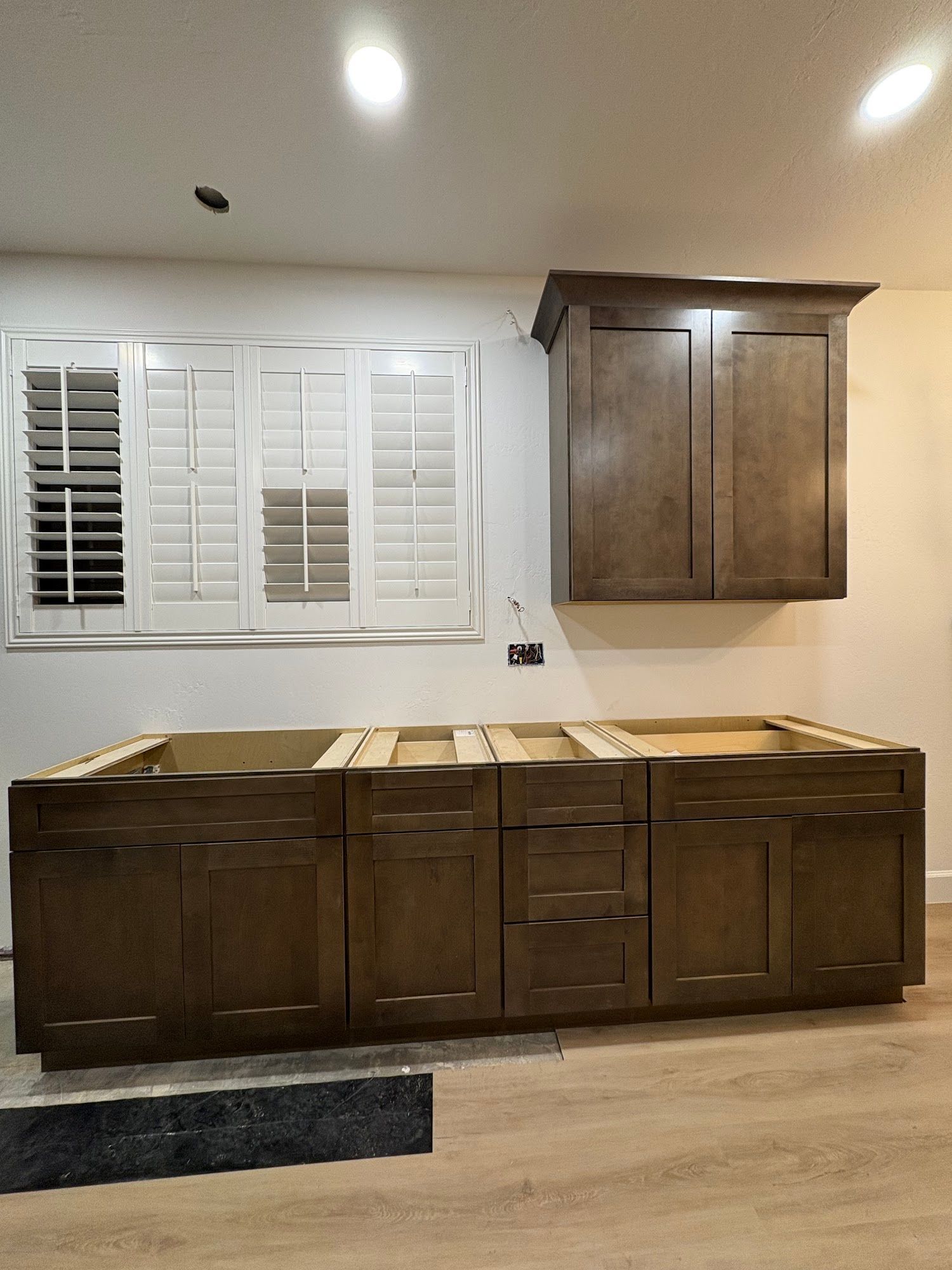 Kitchen cabinets, unfinished installation. Brown base cabinets, matching upper cabinet over window with shutters.