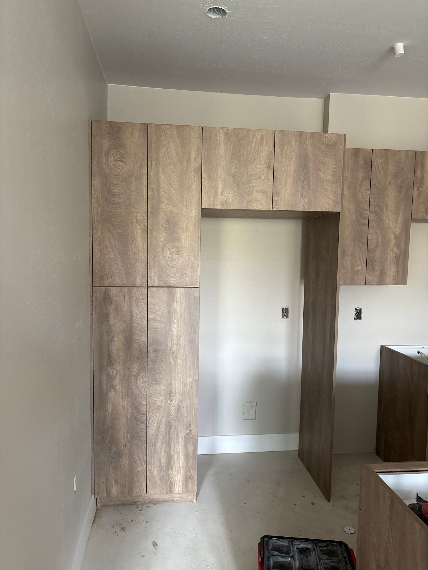 Wooden kitchen cabinets installed against a light beige wall and over a doorway in a room under construction.
