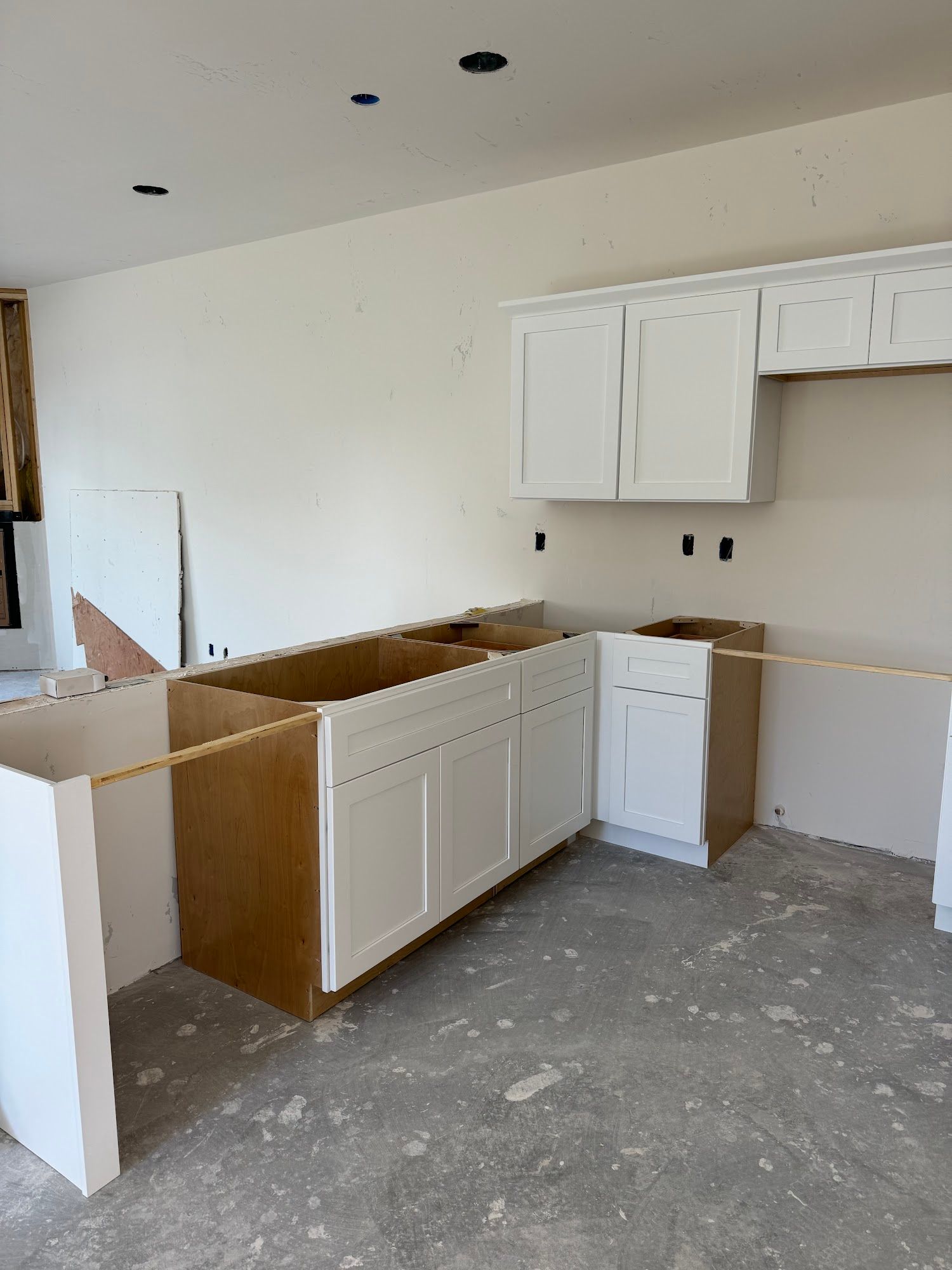 Kitchen cabinets under construction, white cabinets against off-white walls, unfinished flooring.