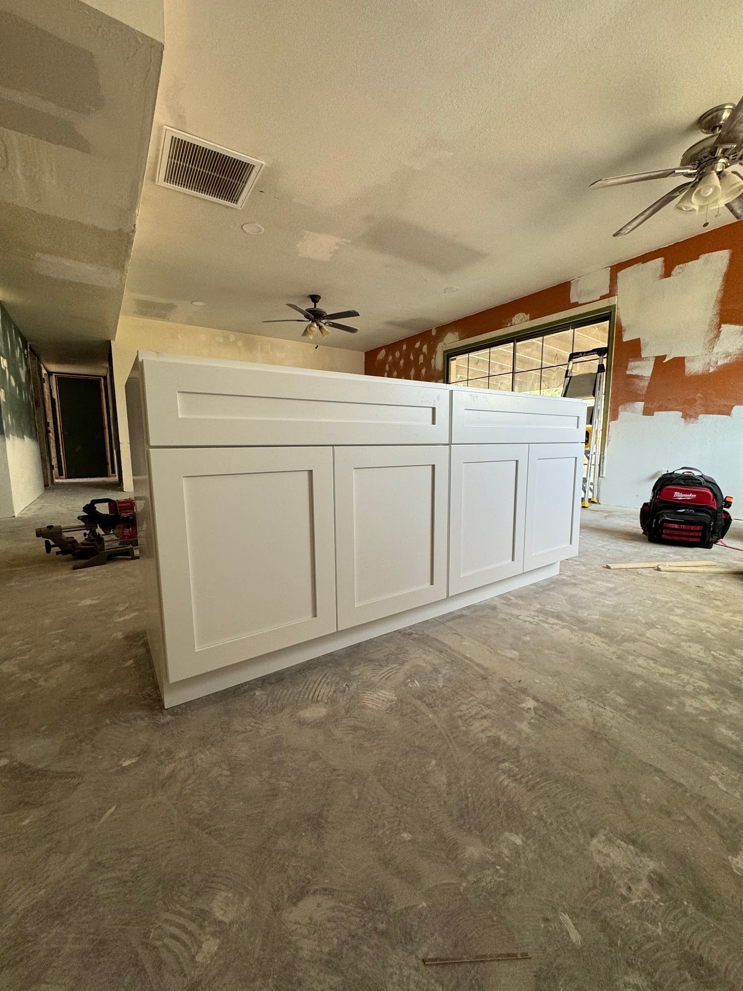 White kitchen cabinets in a room under construction, with exposed walls, ceiling, and flooring.
