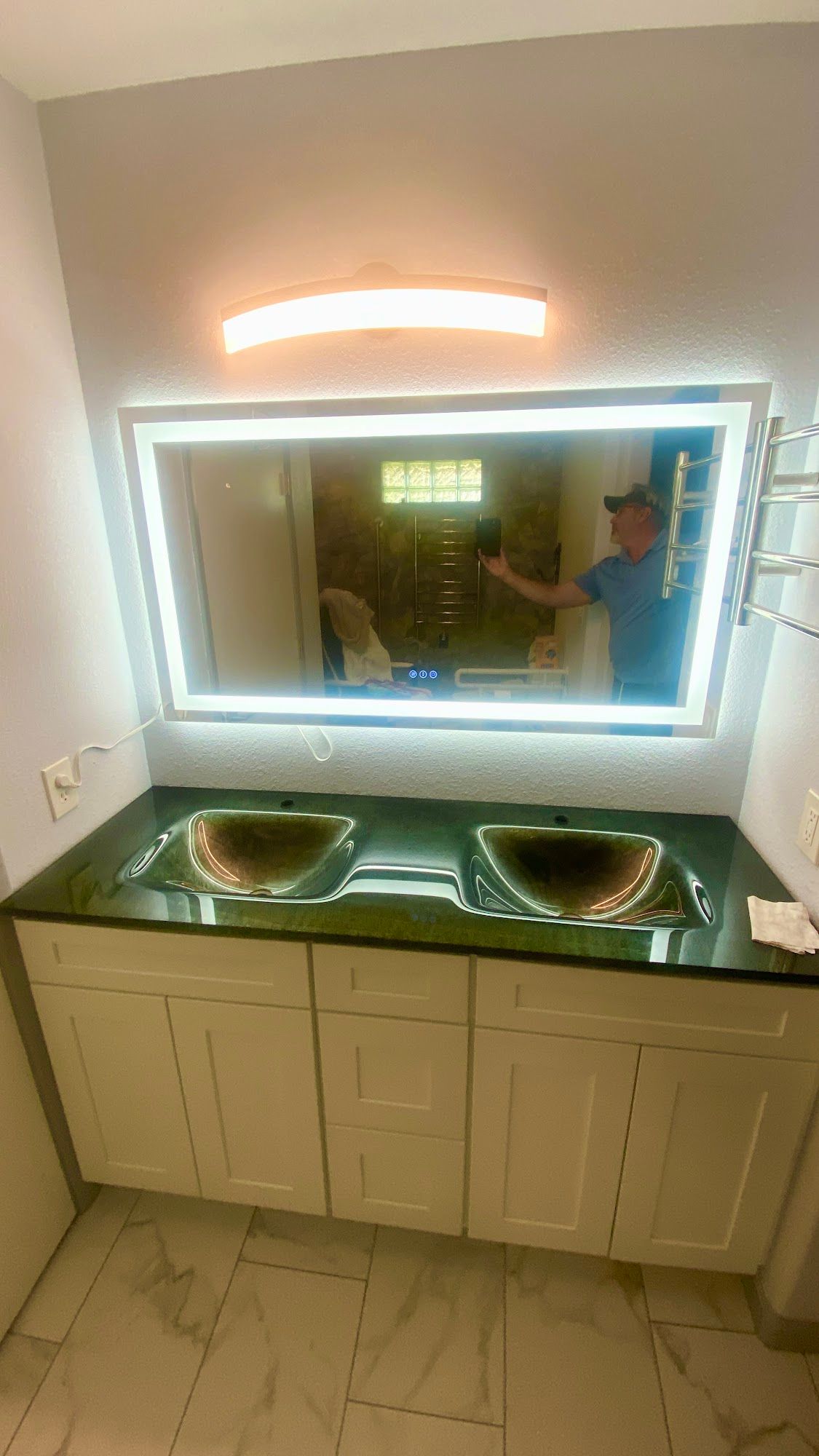 Bathroom with lit mirror, two sinks, white cabinets, person taking photo in reflection.