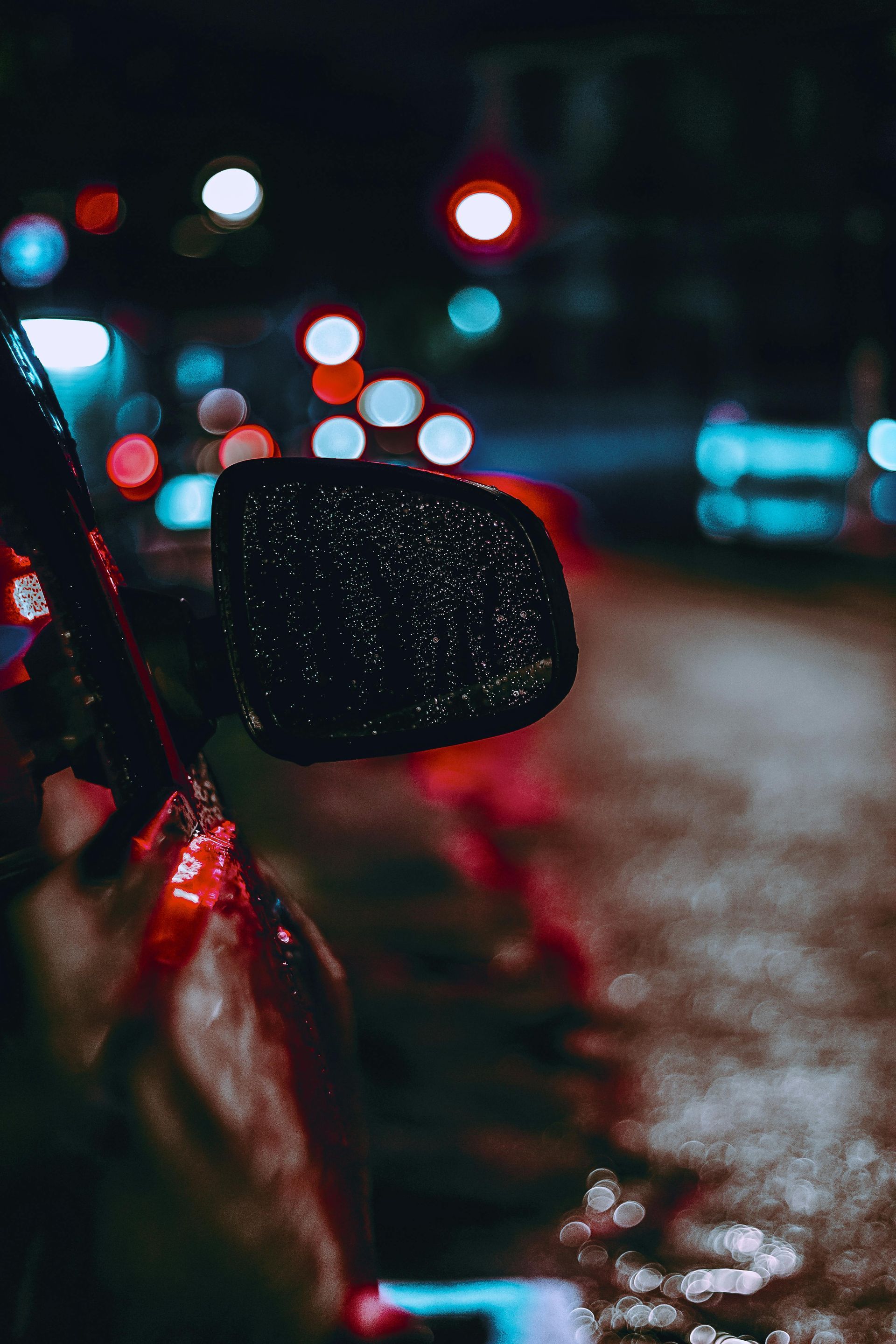 A car side mirror covered in raindrops at night, with blurred, glowing red and blue city lights in the background.