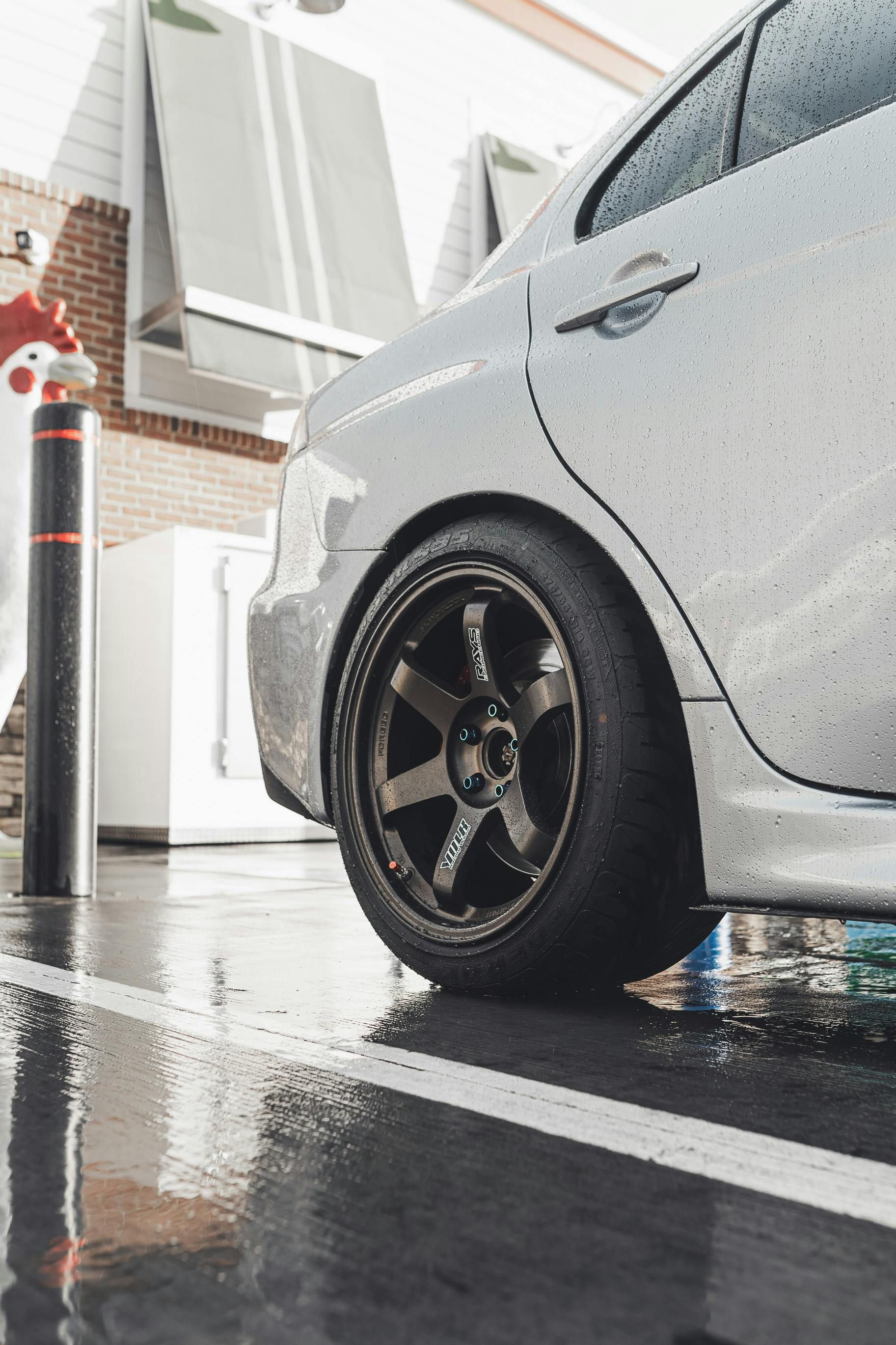 A side-view close-up of a silver car's rear wheel parked on a wet, reflective parking lot surface.