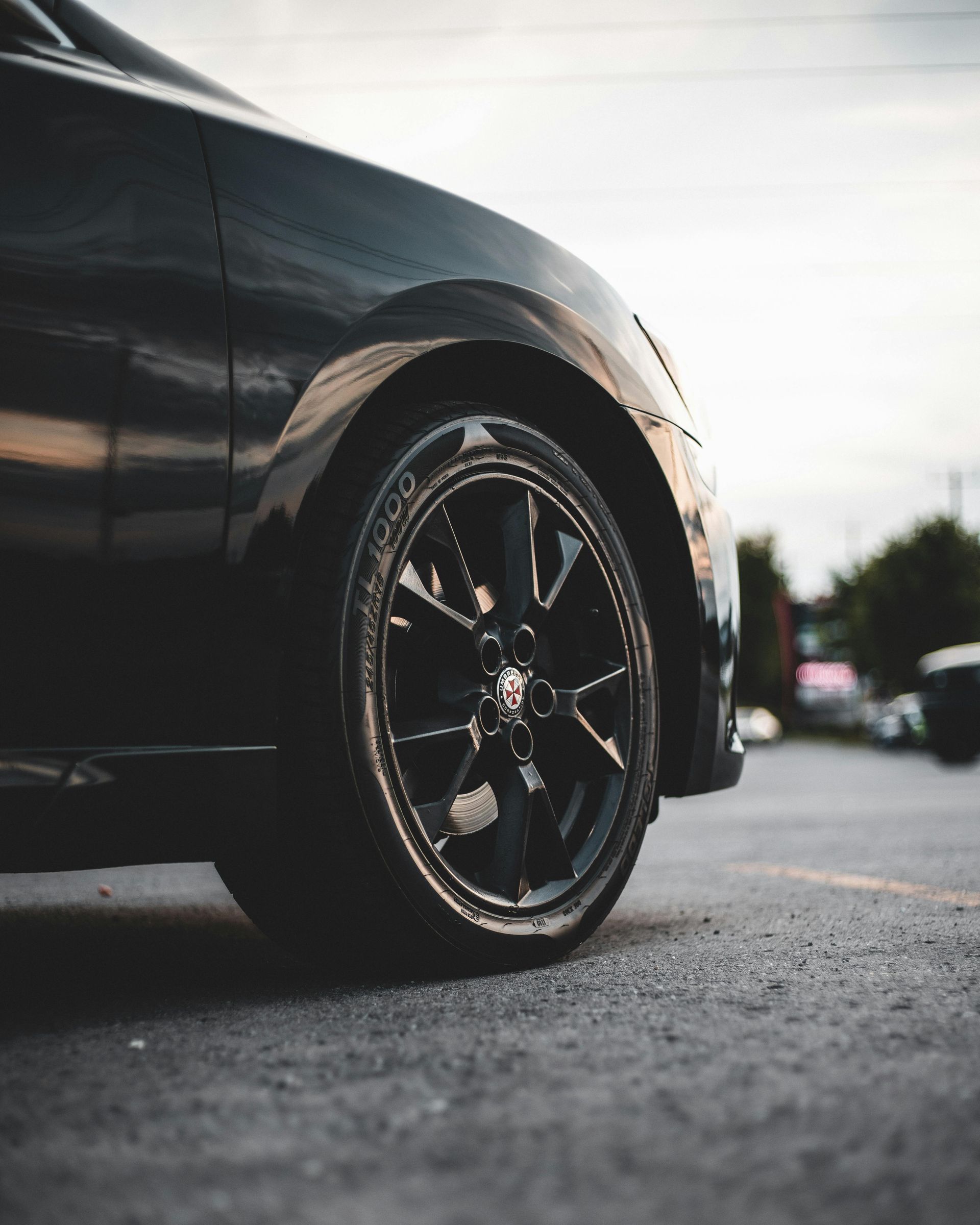 A close-up, low-angle shot of a black car's front tire and wheel parked on an asphalt lot.