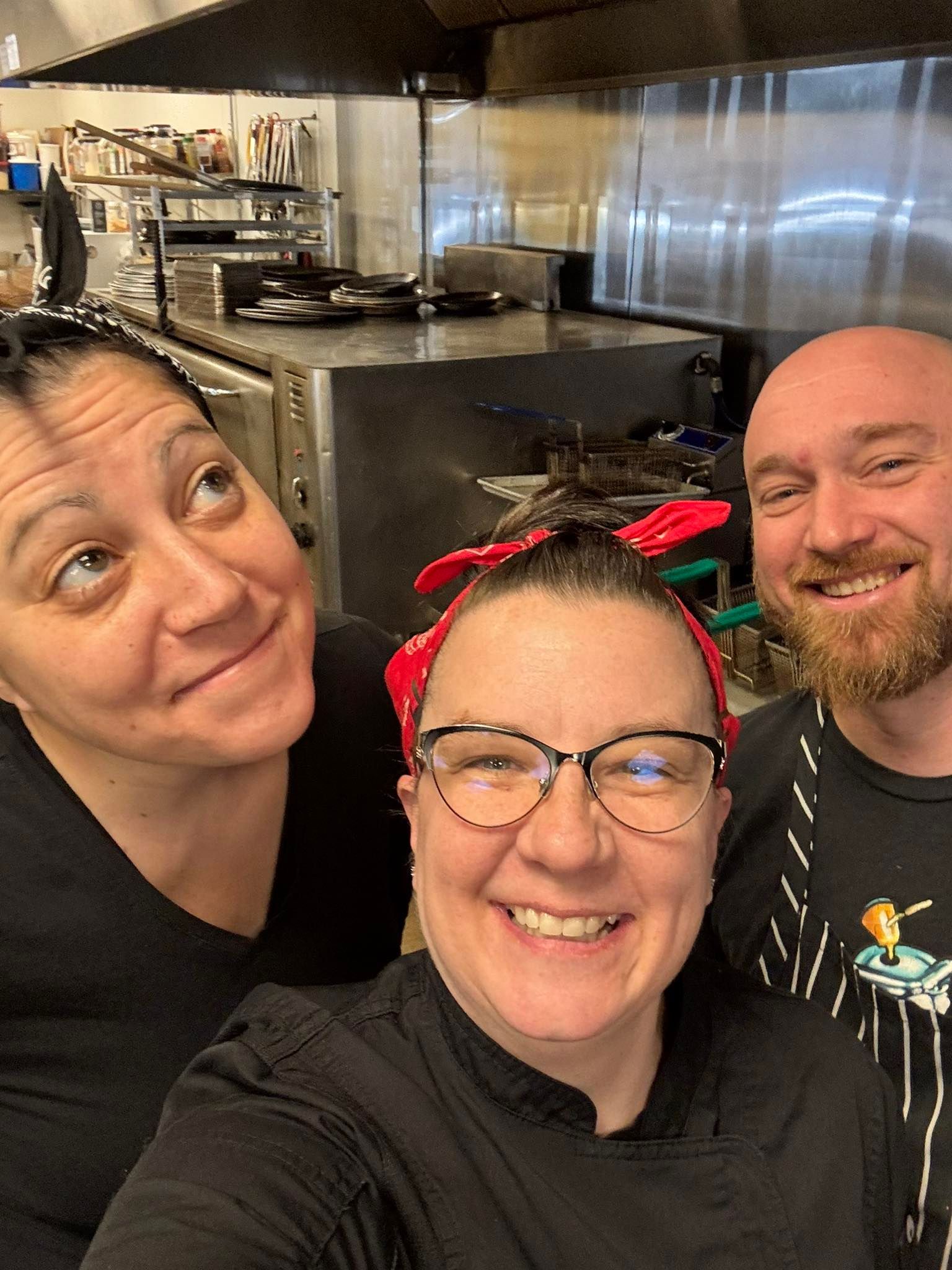 Three people smiling in a commercial kitchen; one looking up, one with glasses and a red headband, and one with a beard.