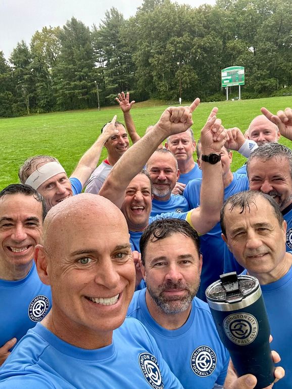 A person wearing a hat and orange shirt, holding a baseball bat on their shoulder on a baseball field.