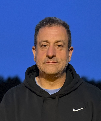 Football coach with arms crossed, looking stern on a sunny field, players in the background.