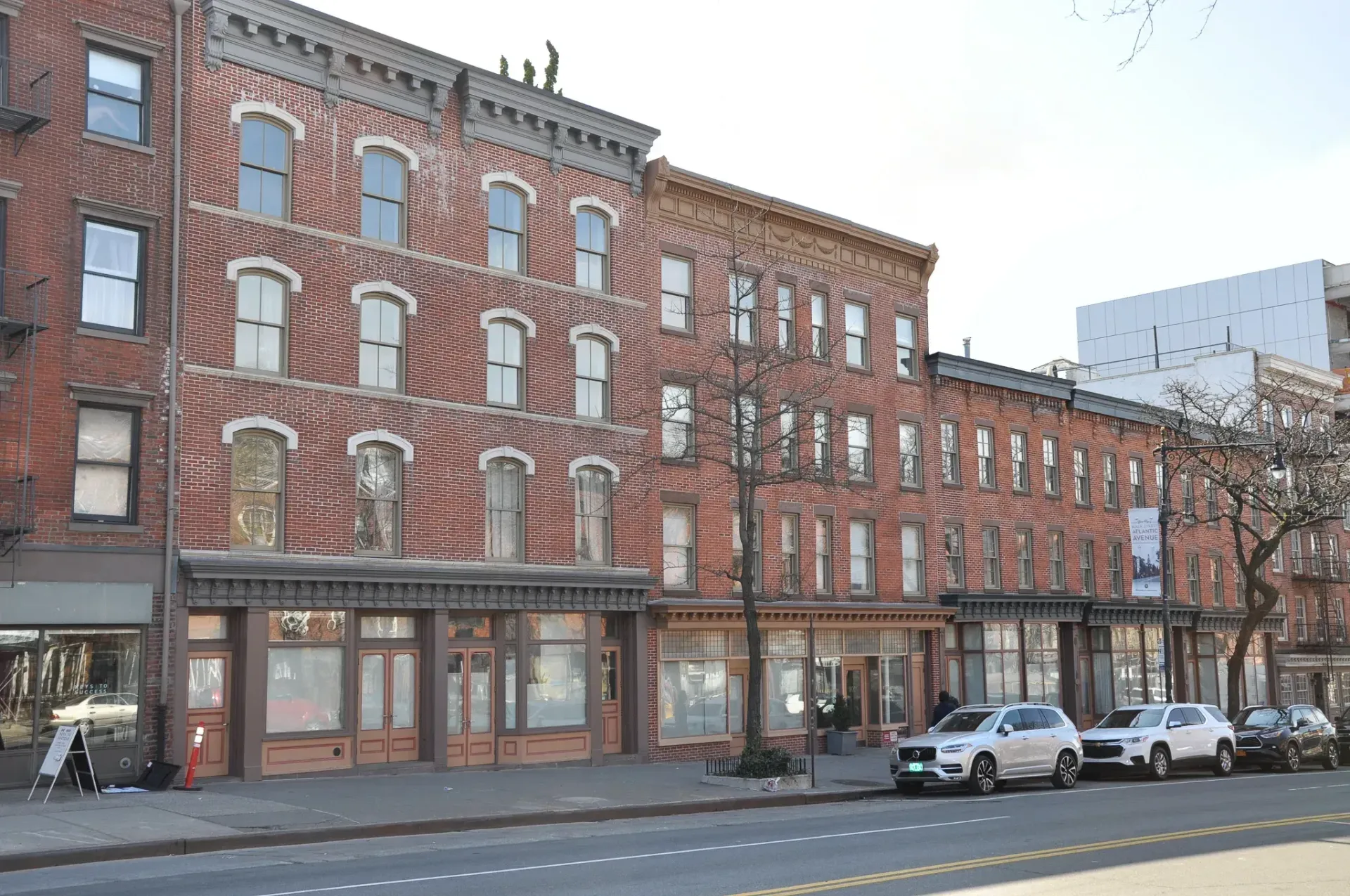 Row of red brick buildings with storefronts and cars parked on the street. Bare trees and a bright sky.