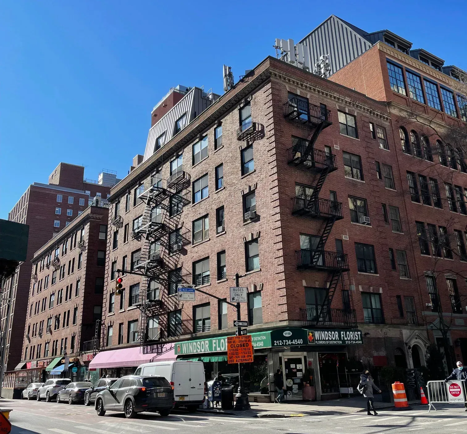 Buildings with brick facades and fire escapes in a city street. Cars parked alongside the street, pedestrians walking.
