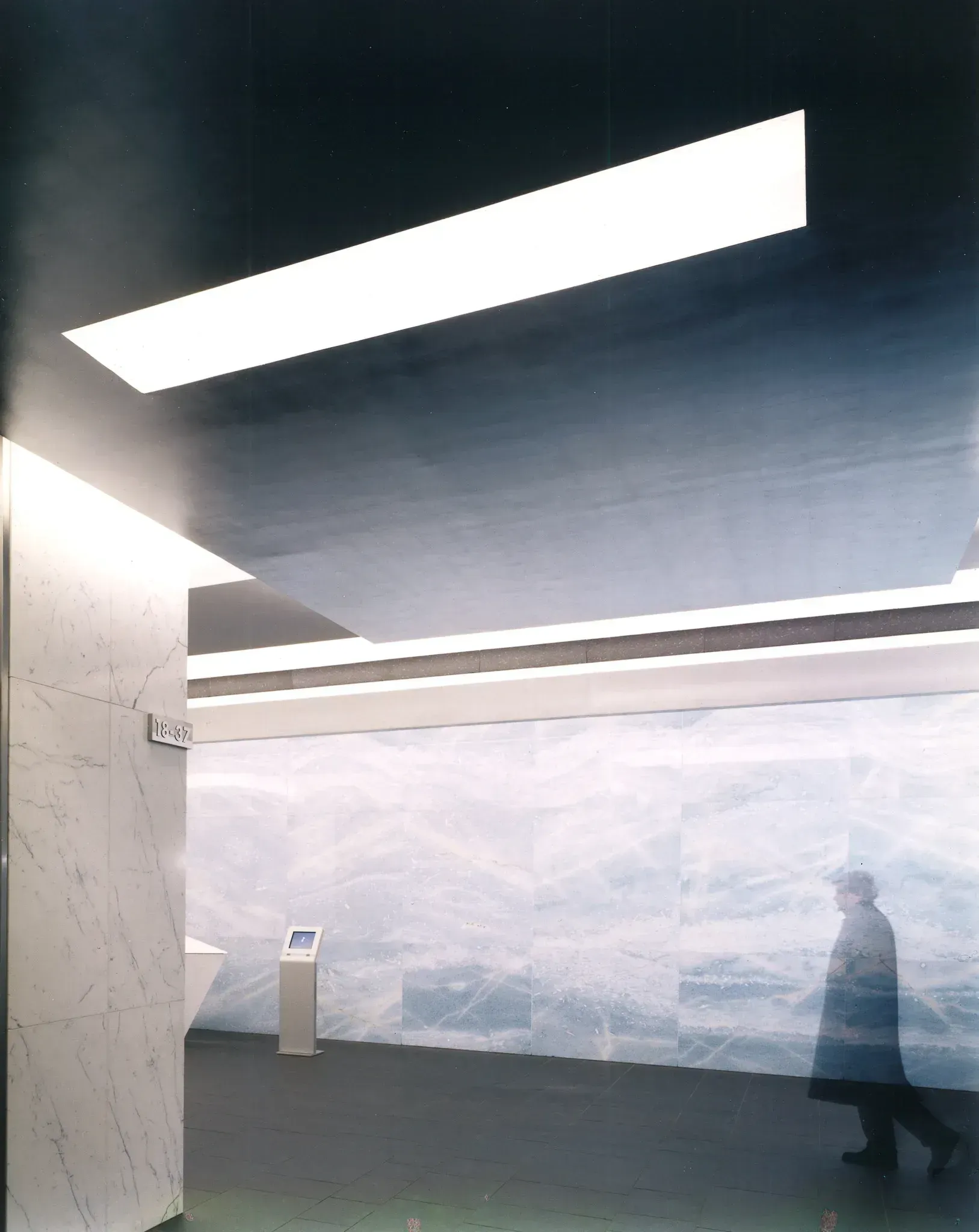 A person in a coat walks through a modern, minimalist space with white walls and bright overhead lighting.