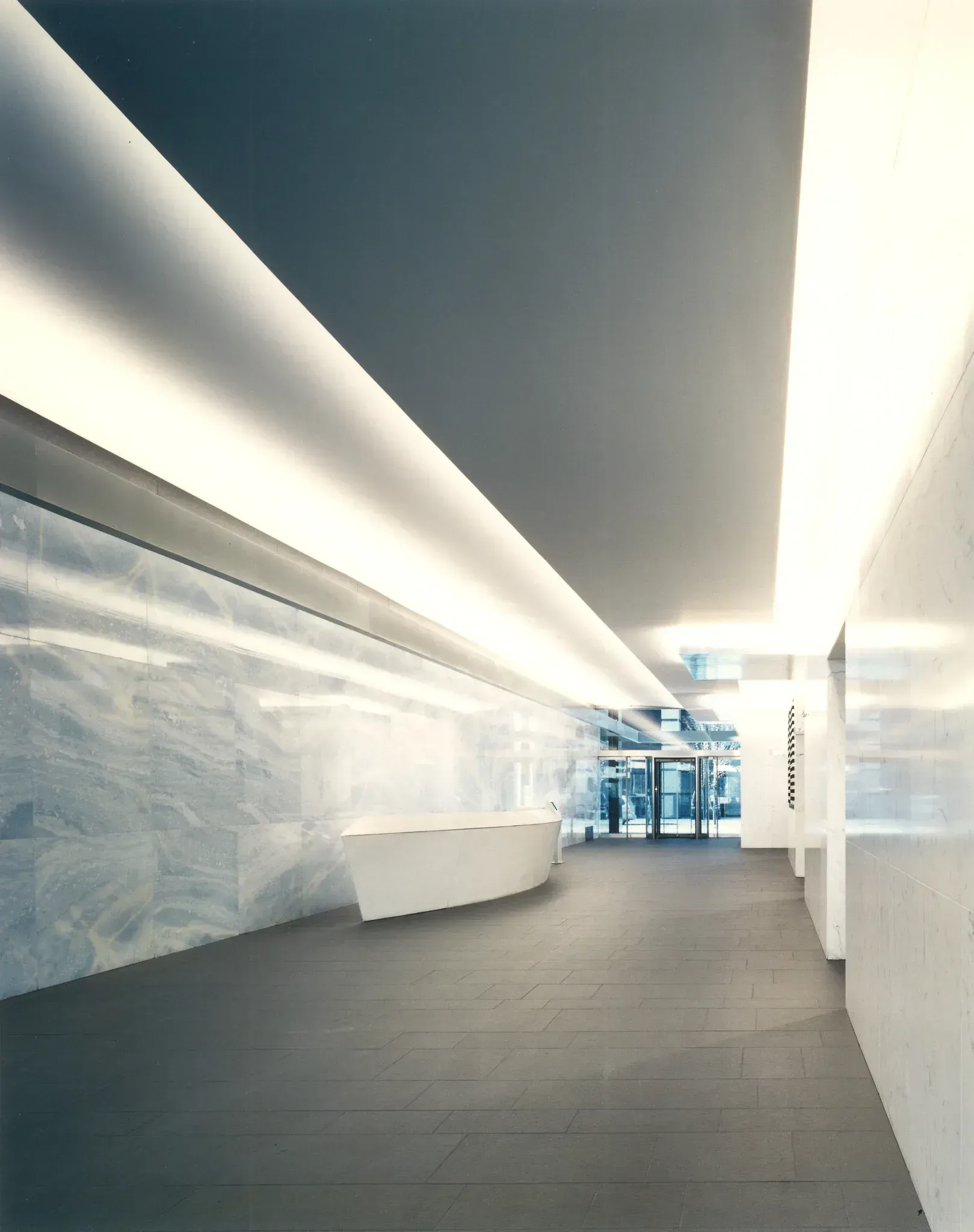 Interior hallway with long, white walls, a curved white reception desk, and overhead lighting.