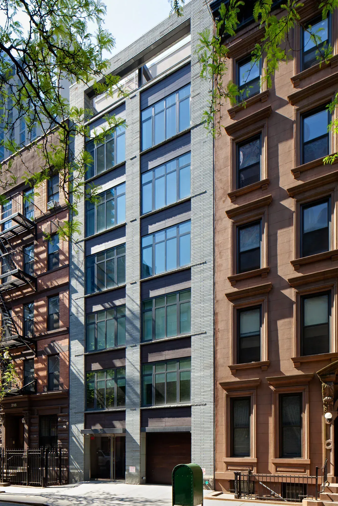 A tall, modern building with many windows stands between two older brownstone buildings in a city setting.