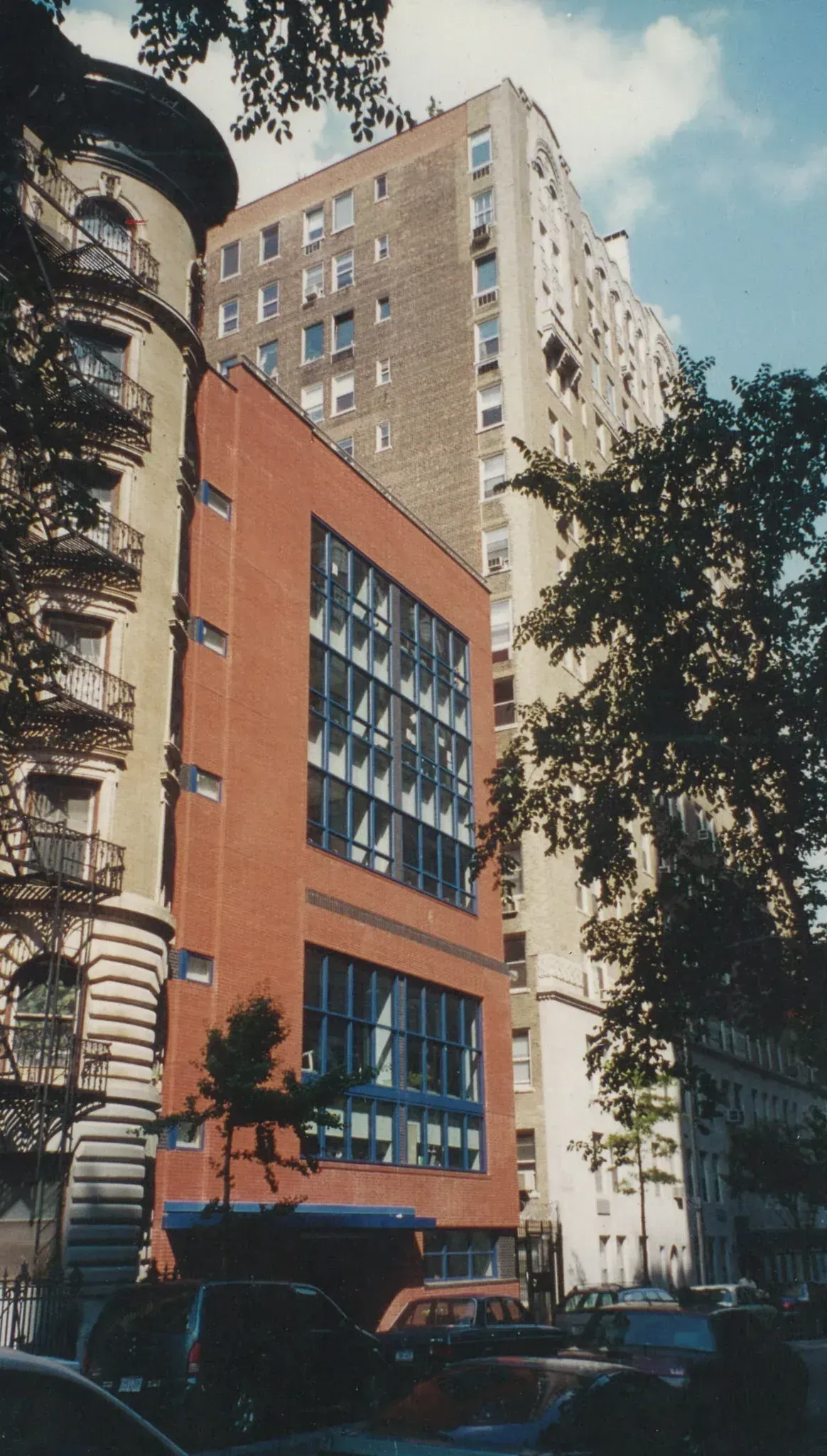 A brick building with large windows stands between taller apartment buildings on a city street.