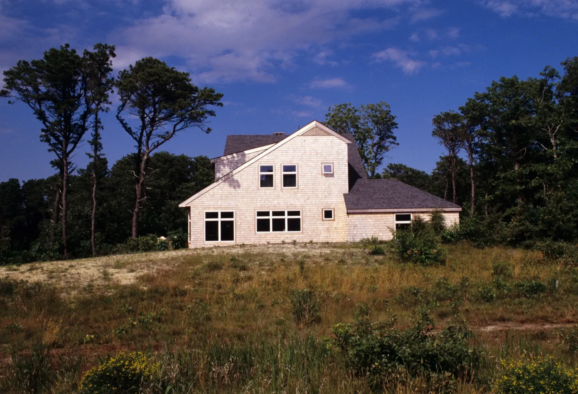 House under construction on a grassy hill, surrounded by trees. The sky is blue with some clouds.